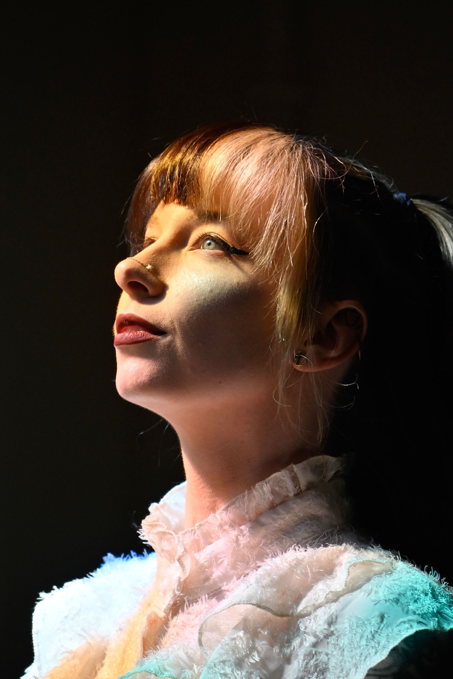 A person in a white ruffled top looks upward with closed eyes as dramatic lighting illuminates their face from the side.