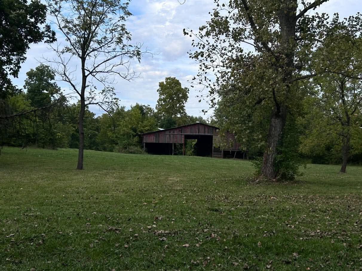 A rustic wooden barn sits in a lush green meadow surrounded by tall trees under a partly cloudy sky.