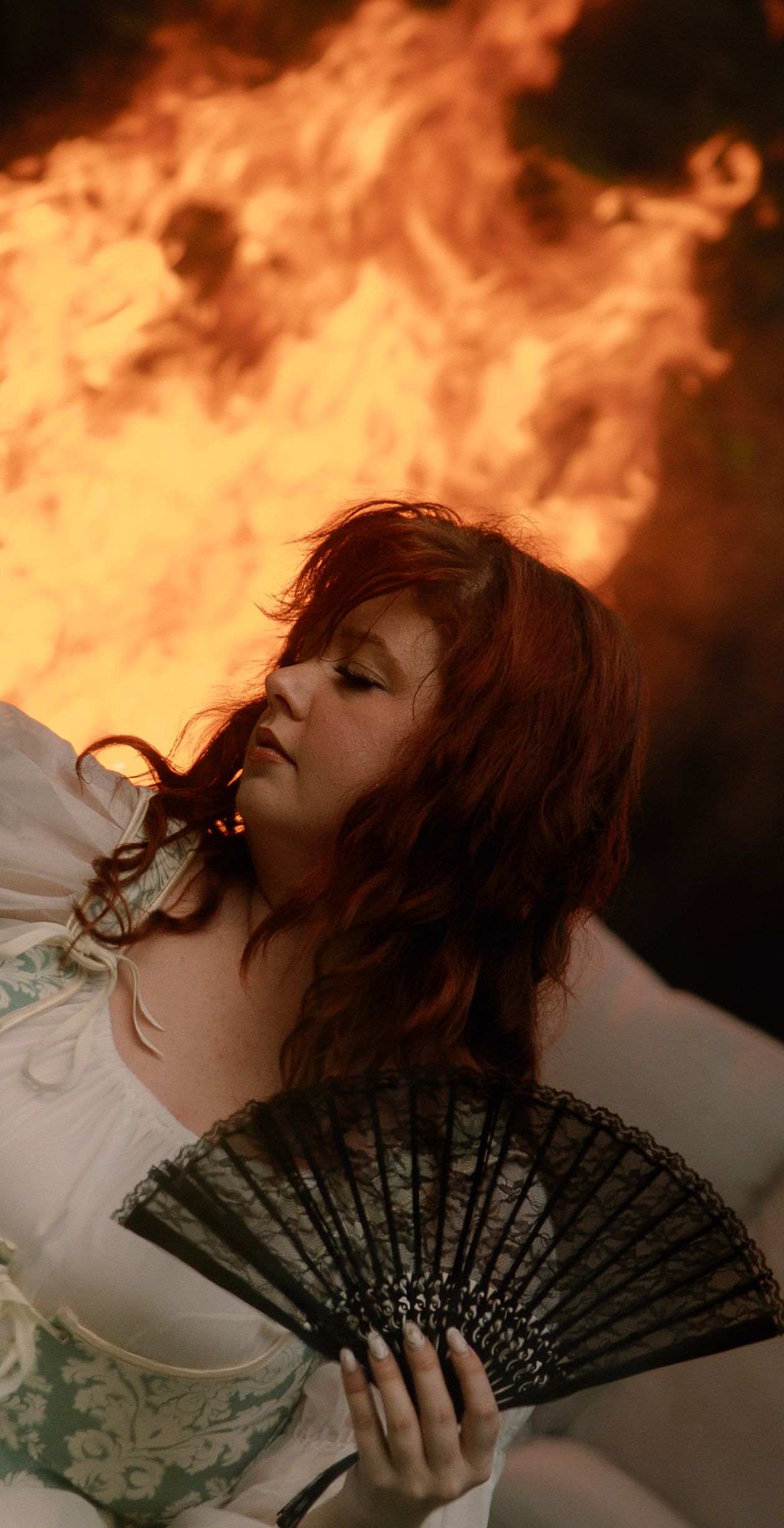 A red-haired woman in a white Victorian dress holds a black lace fan with dramatic flames burning behind her.