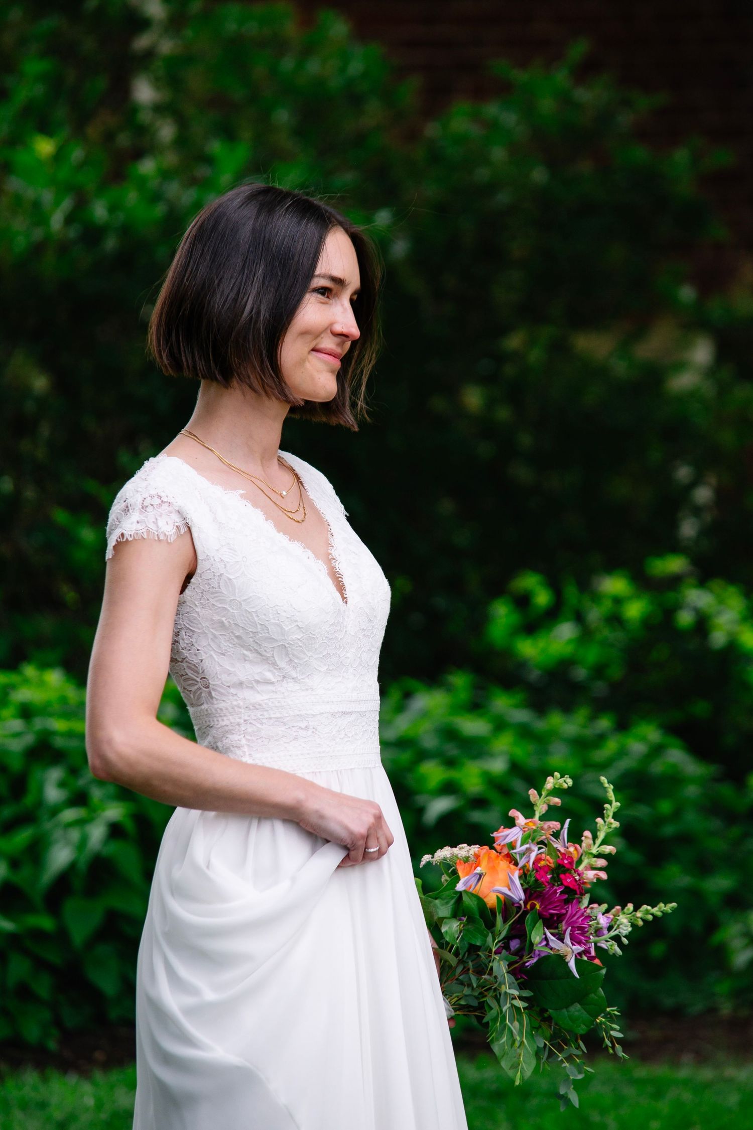 Bride in white lace cap-sleeve wedding dress holding colorful bouquet, smiling in lush green garden.
