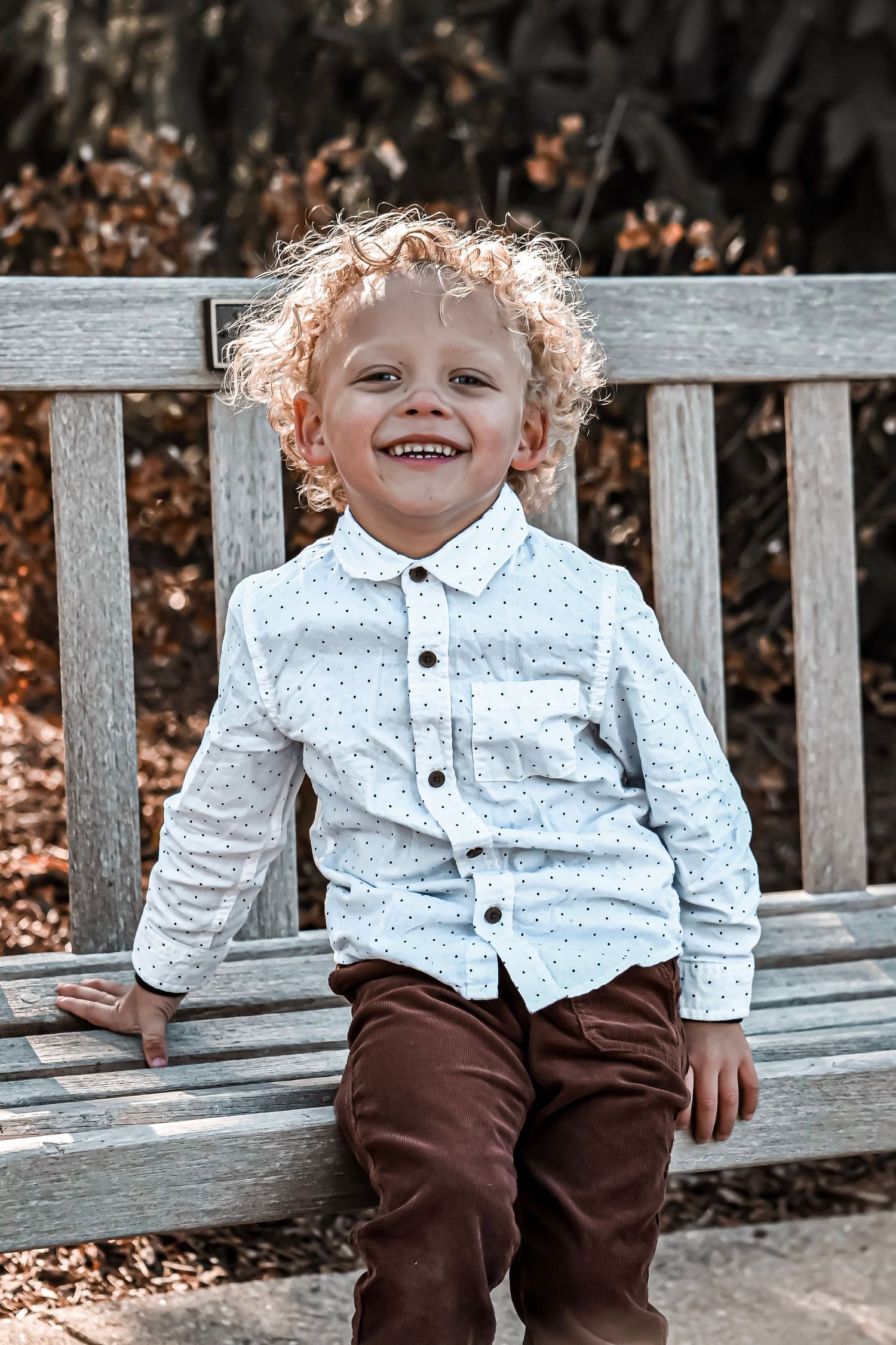 Child in white button-down shirt and brown pants sits smiling on wooden bench outdoors.