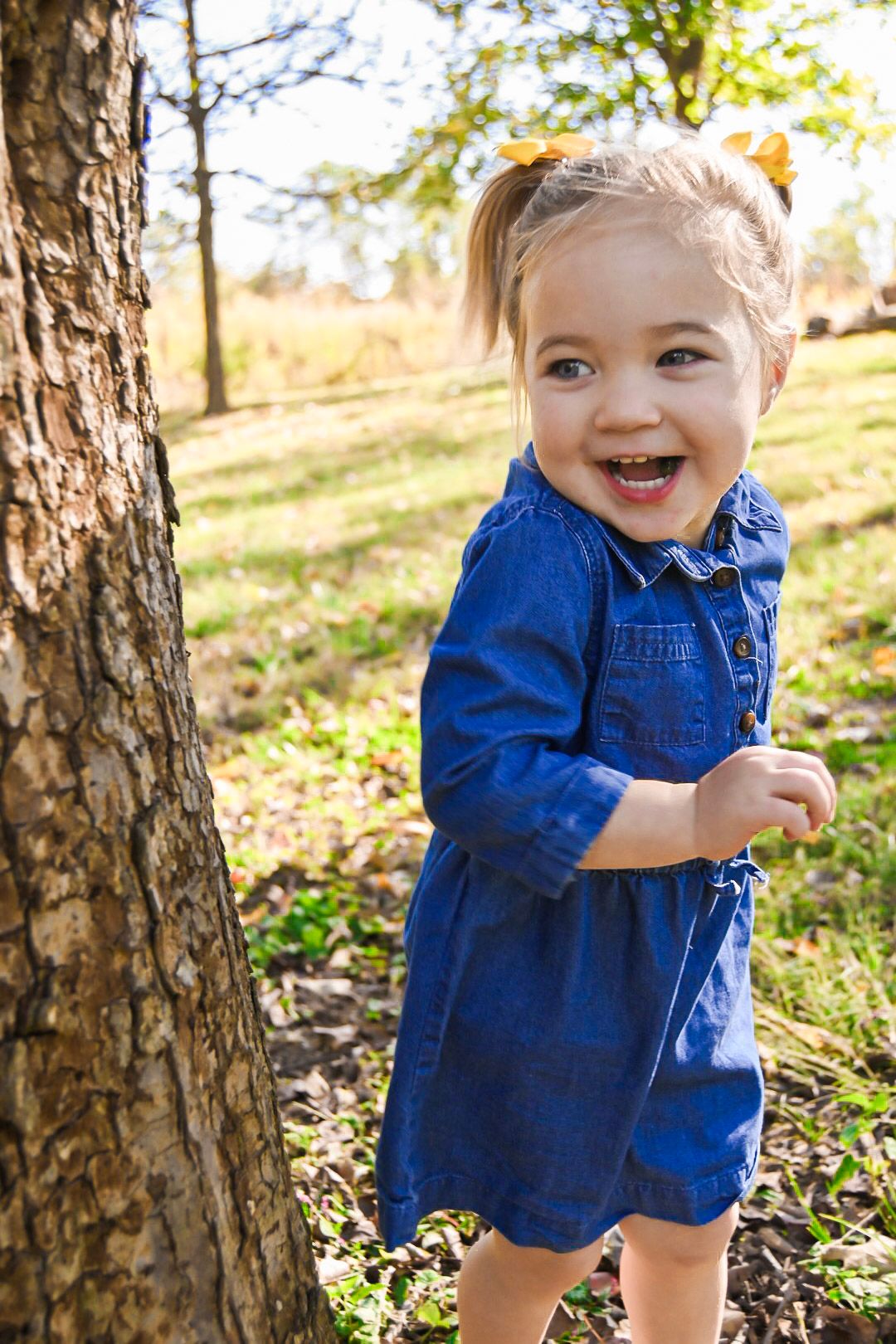 Toddler in blue denim dress playing among trees in autumn sunlight.