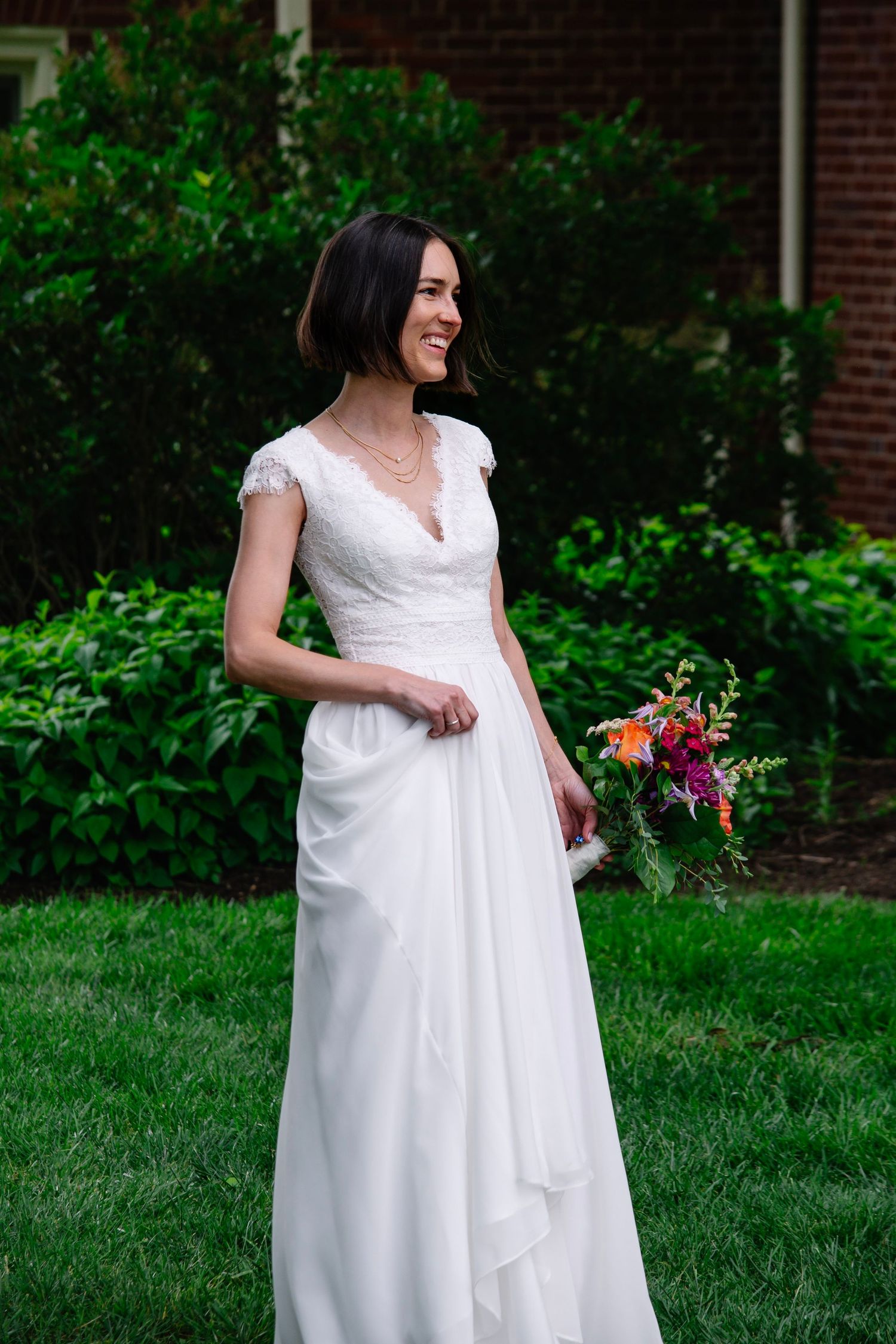 Smiling bride in white lace cap-sleeve wedding gown holding colorful bouquet, standing on green lawn.