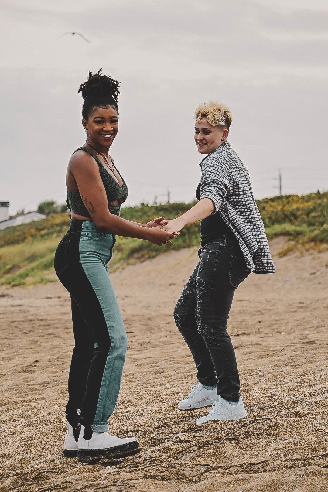 A couple dances playfully together on a sandy beach with coastal vegetation in the background.