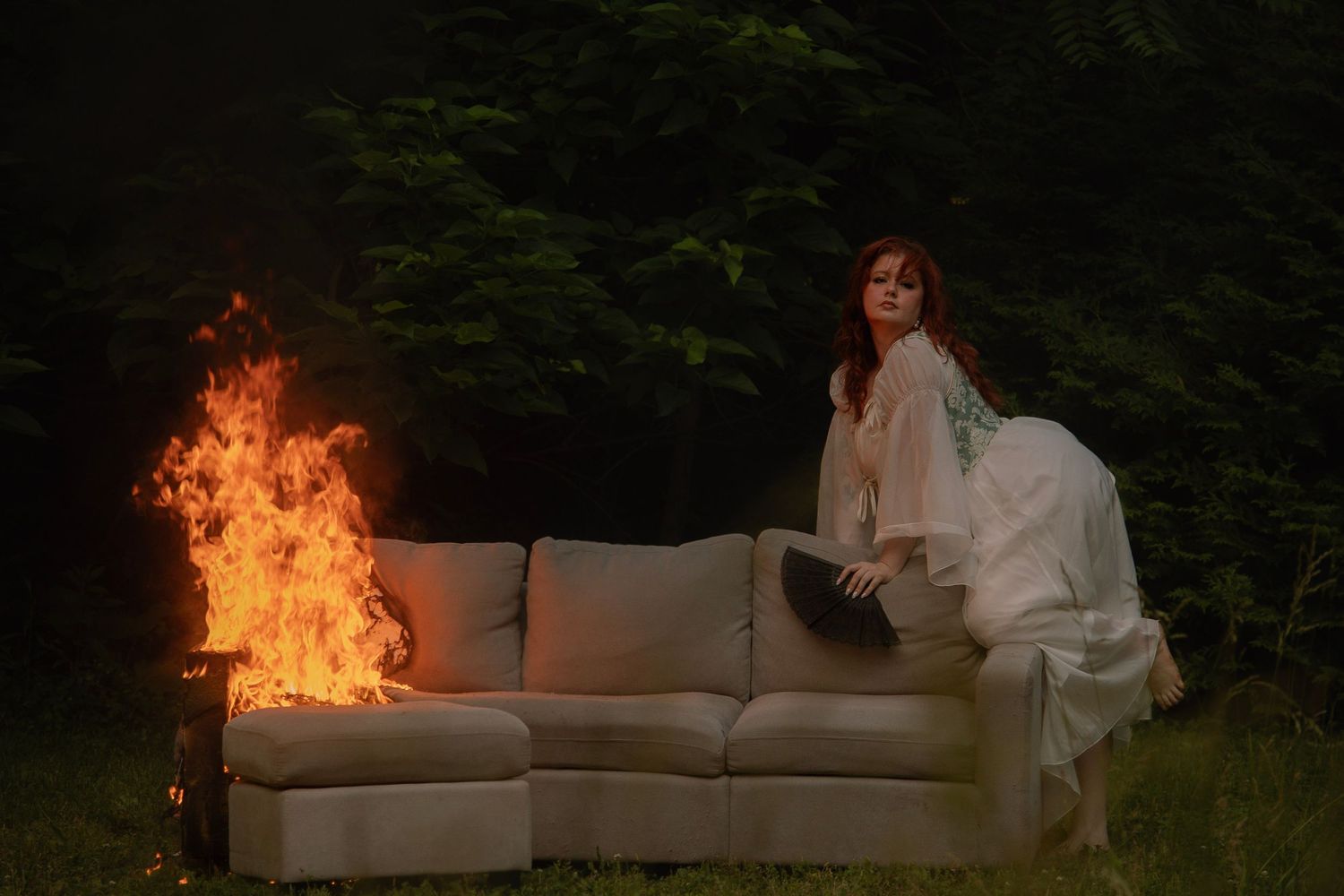 Woman in white gown sits on cream sofa outdoors as flames burn beside her against dark forest backdrop.