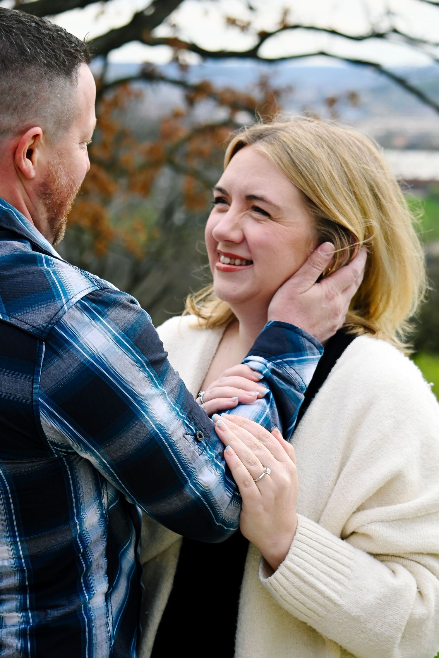 A smiling blonde woman gazes at a man in a blue plaid shirt as he gently touches her face outdoors.