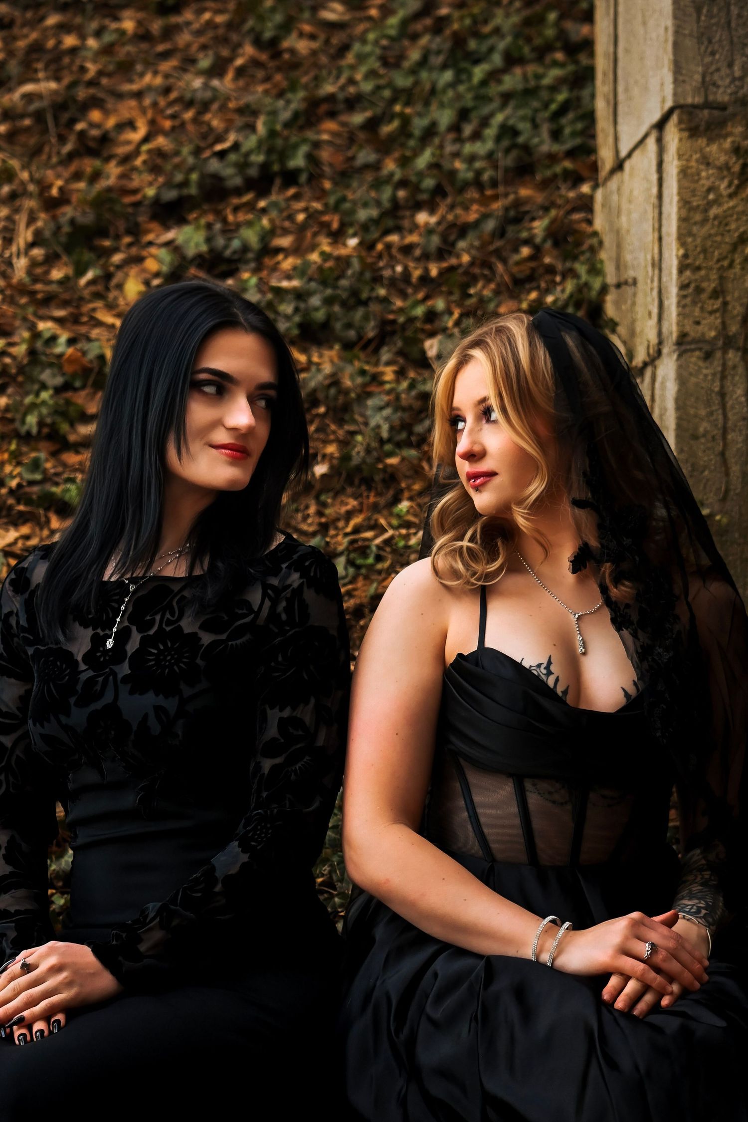 Two people in black dresses sit close together on stone steps surrounded by autumn foliage.