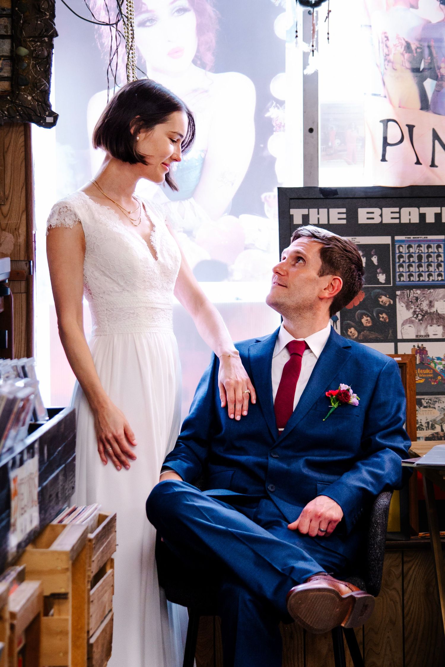 Bride in white gown and groom in blue suit share a romantic moment in a vintage record shop with Beatles poster.