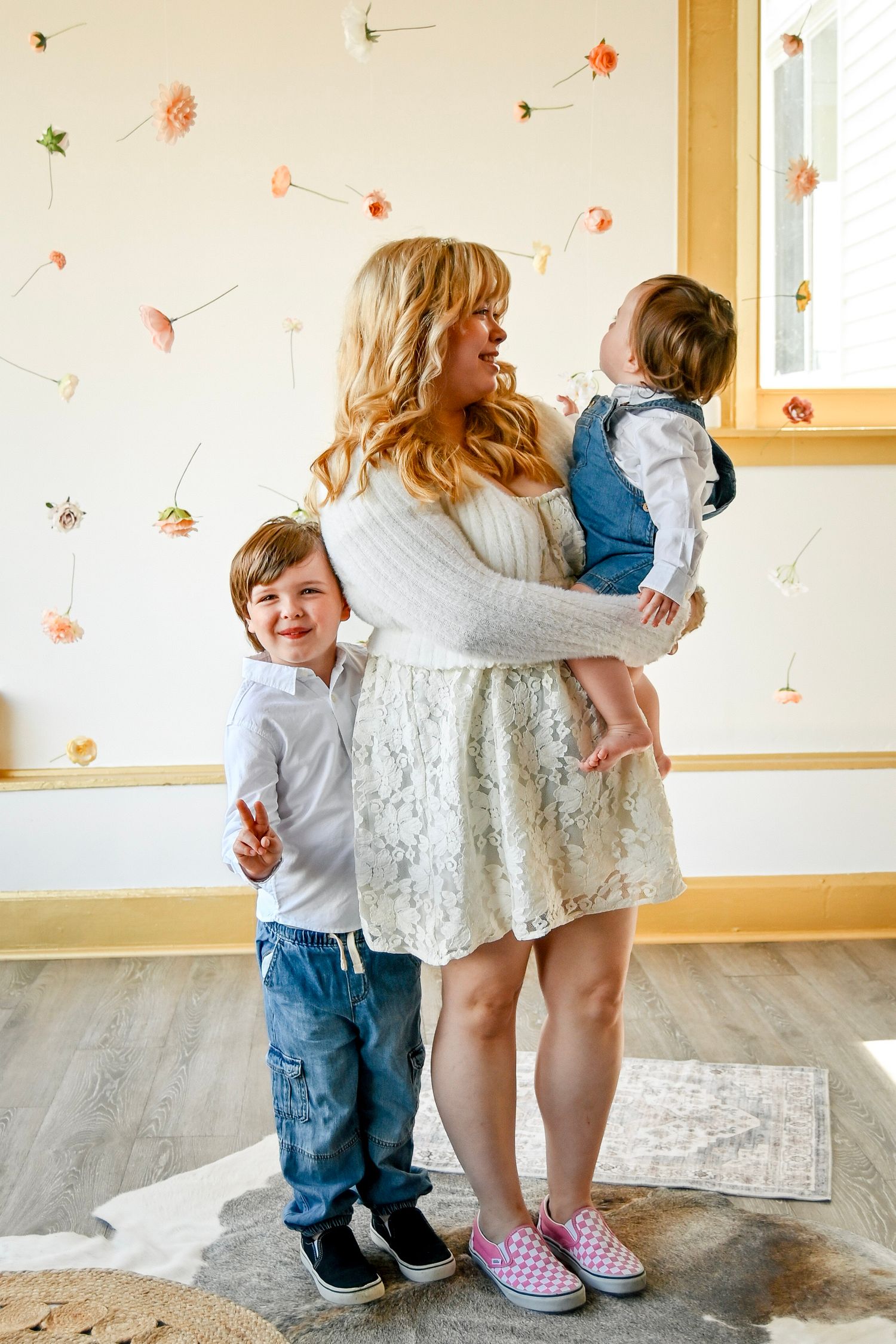 A blonde mother in a white lace dress holds a baby while her young son stands beside her in a floral decorated room.