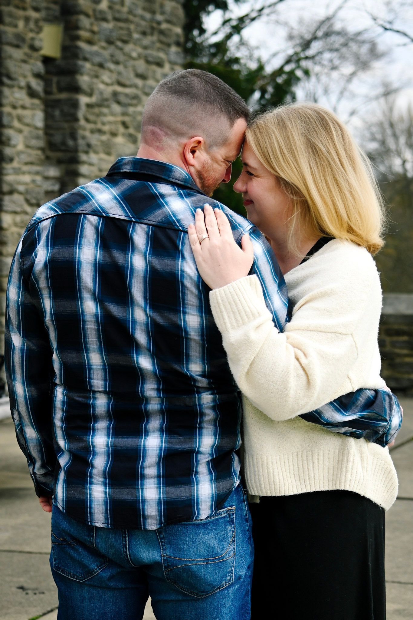 A couple sharing an intimate moment, touching foreheads outdoors near a stone wall, woman in white sweater, man in plaid shirt.