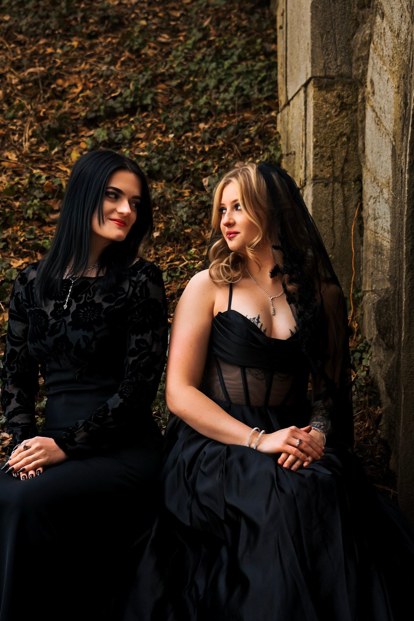 Two women in black dresses pose together in an outdoor wooded setting with autumn leaves on the ground.