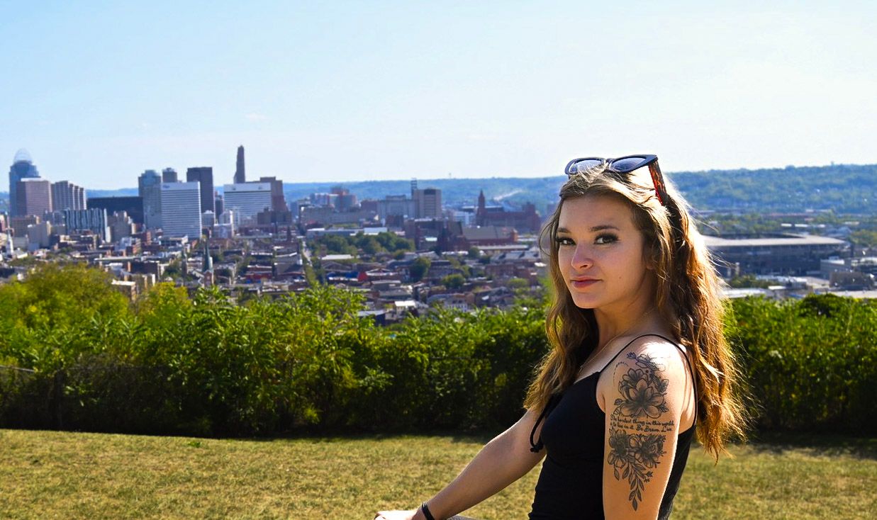 Woman with tattoo overlooking city skyline from hilltop viewpoint.