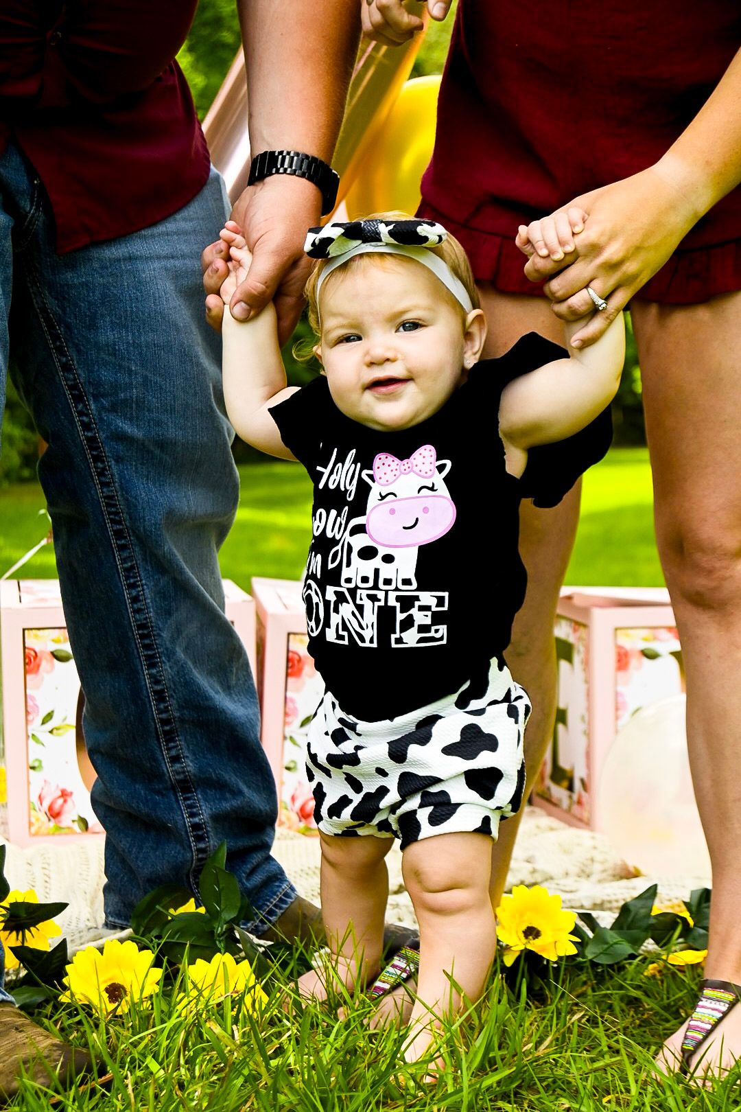 Baby wearing cow print outfit sits on grass near decorative blocks.
