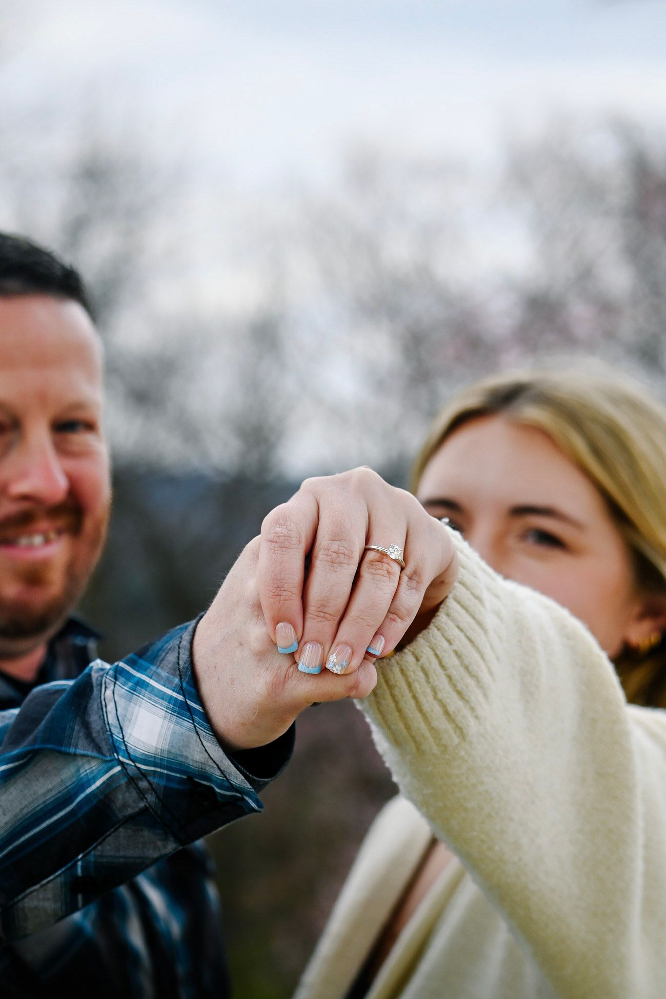 Happy couple holding hands showing engagement ring outdoors with bare trees in background.