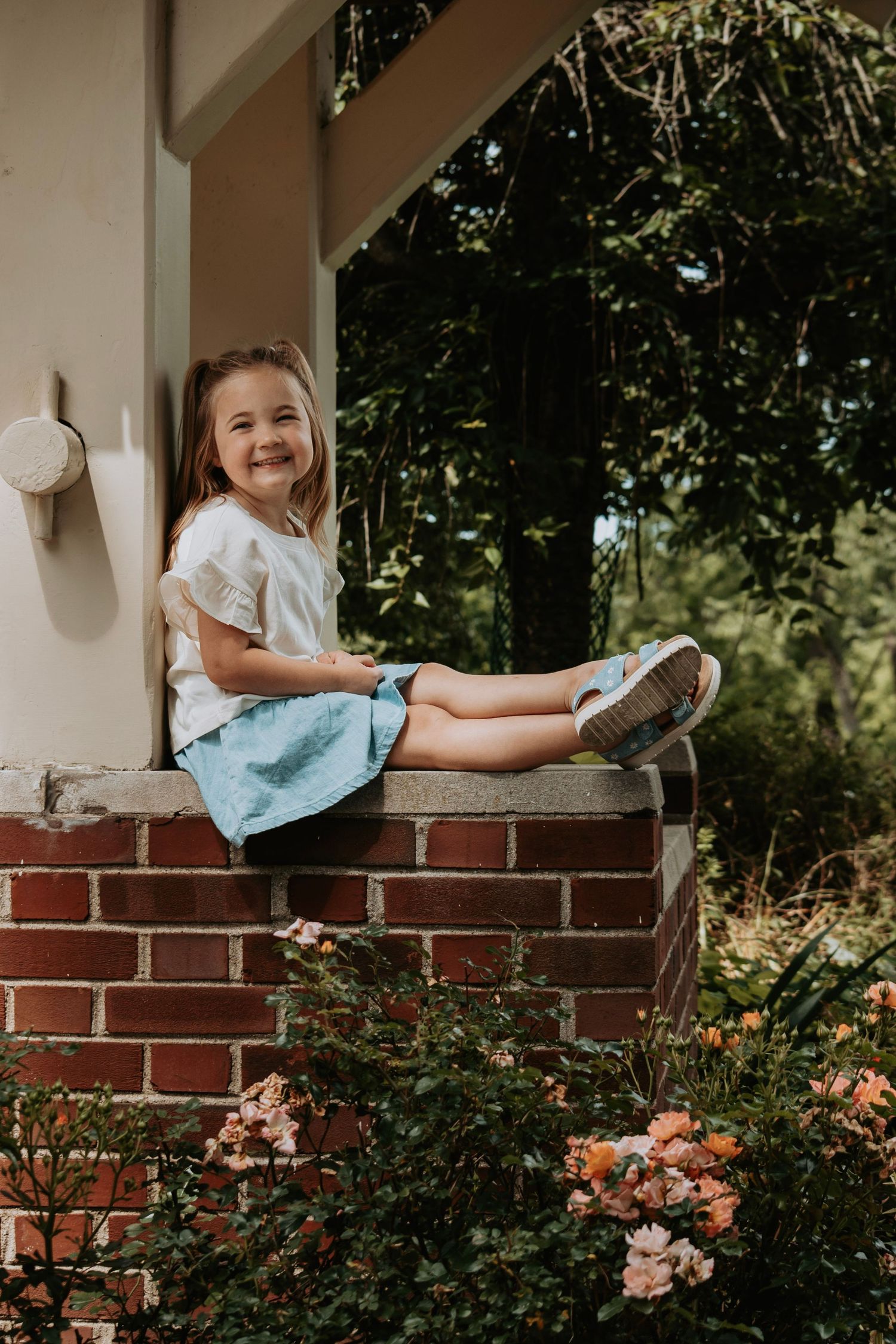A smiling young girl in a white top and blue skirt sits on a brick porch wall surrounded by blooming roses.
