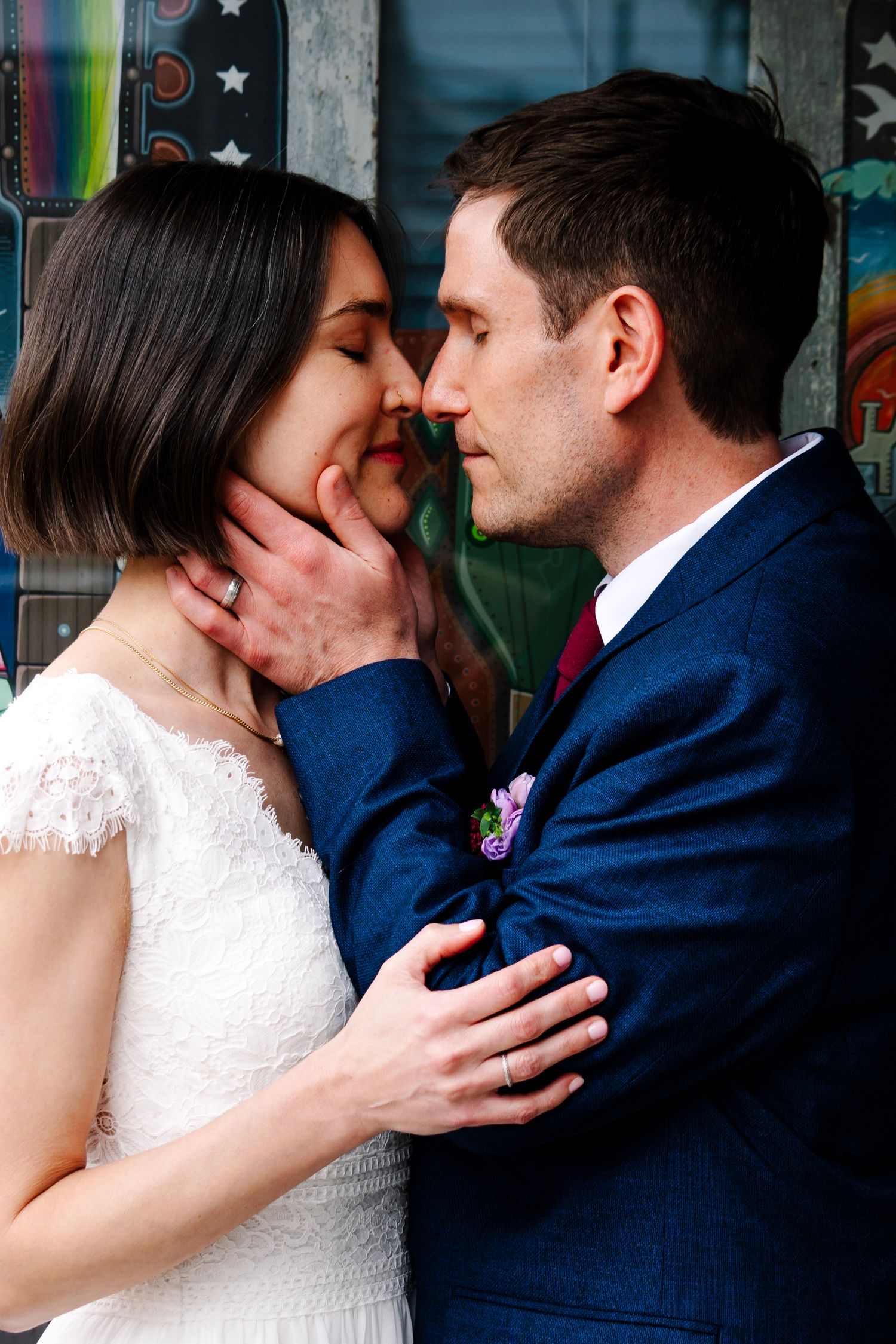 Bride and groom share a romantic moment, nearly kissing, with groom in blue suit and bride in white lace dress.