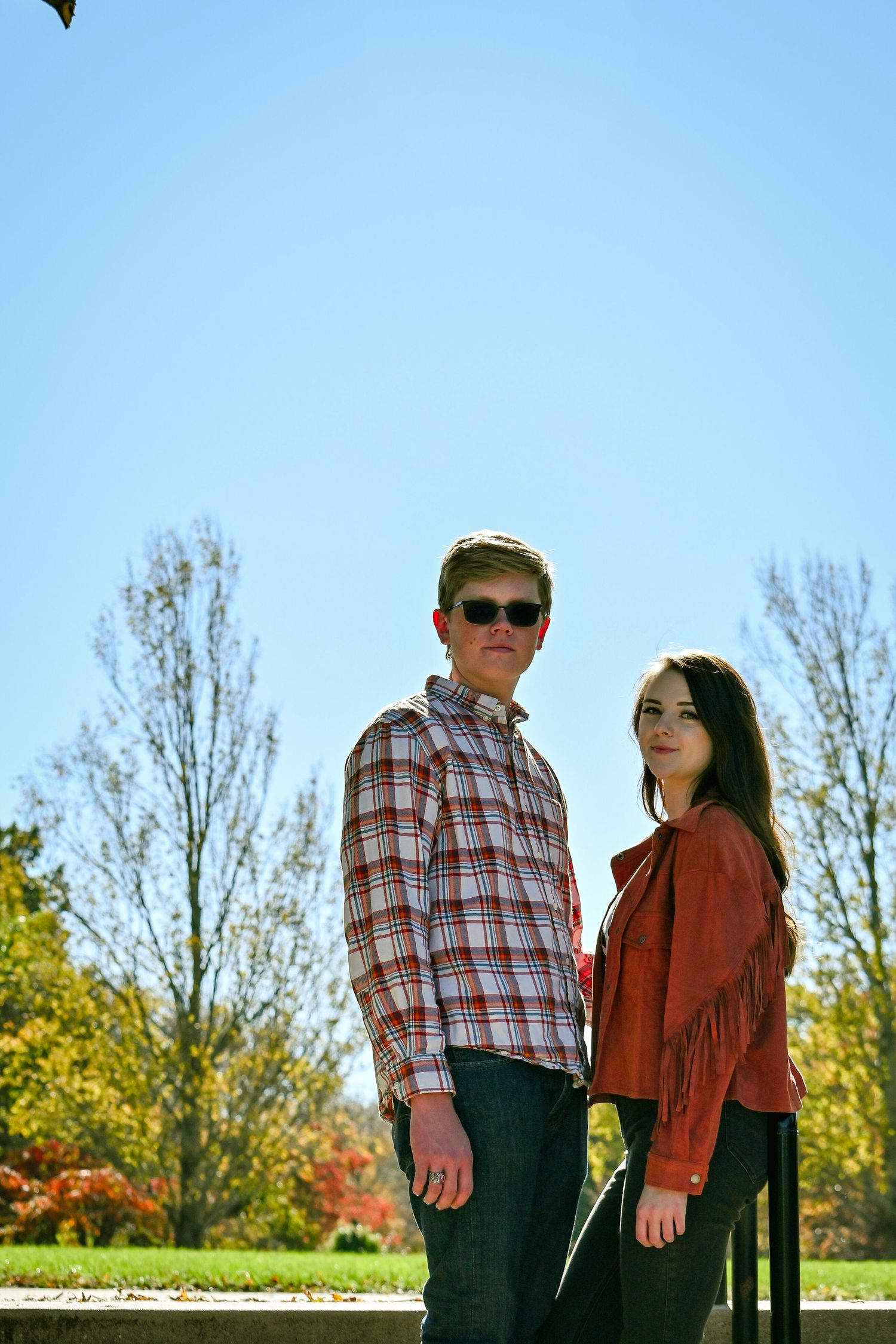 A couple stands outdoors on a sunny autumn day wearing casual attire including a plaid shirt and fringed jacket.
