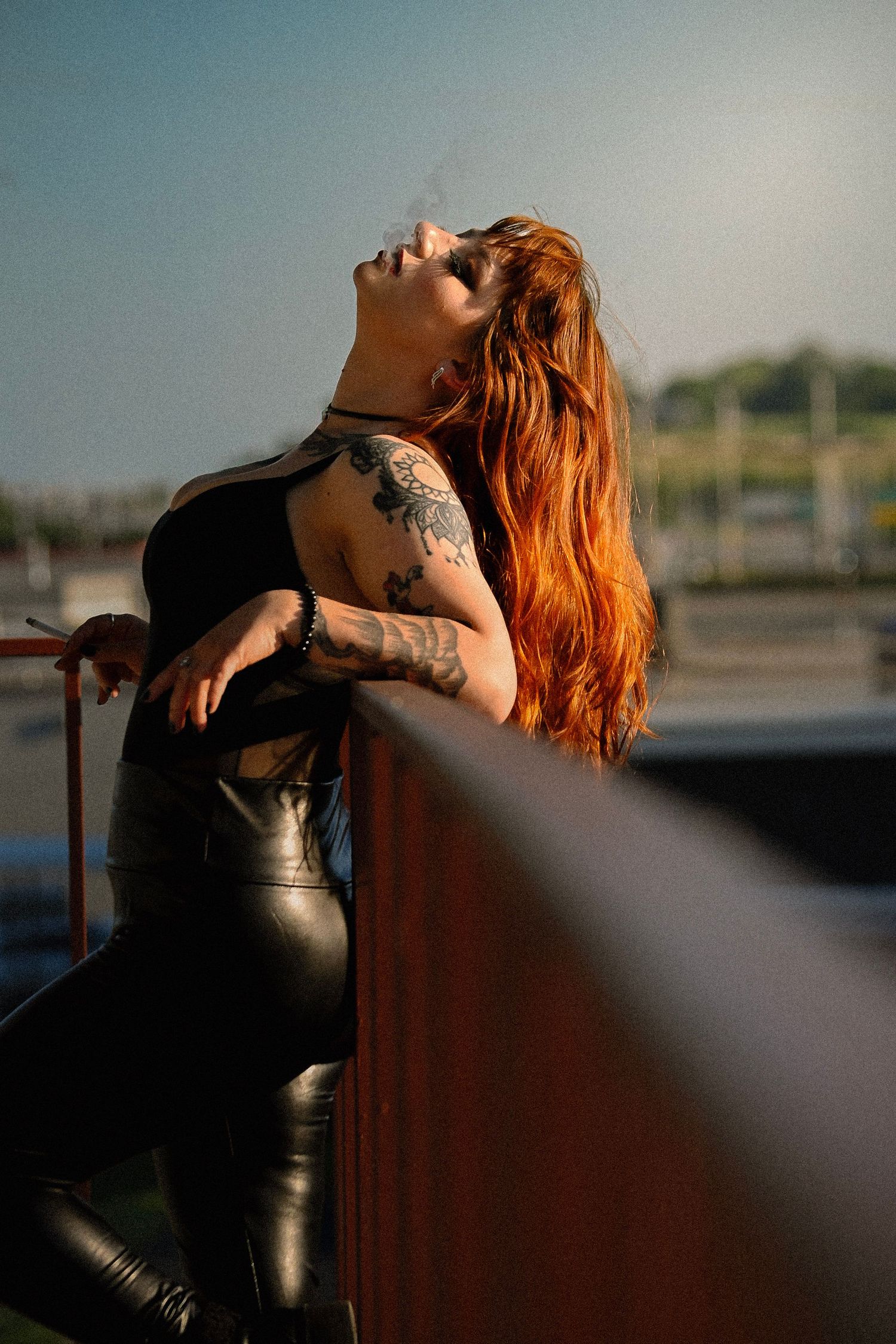 Red-haired woman with tattoos in black leather outfit leaning on rusty railing outdoors in golden hour light.