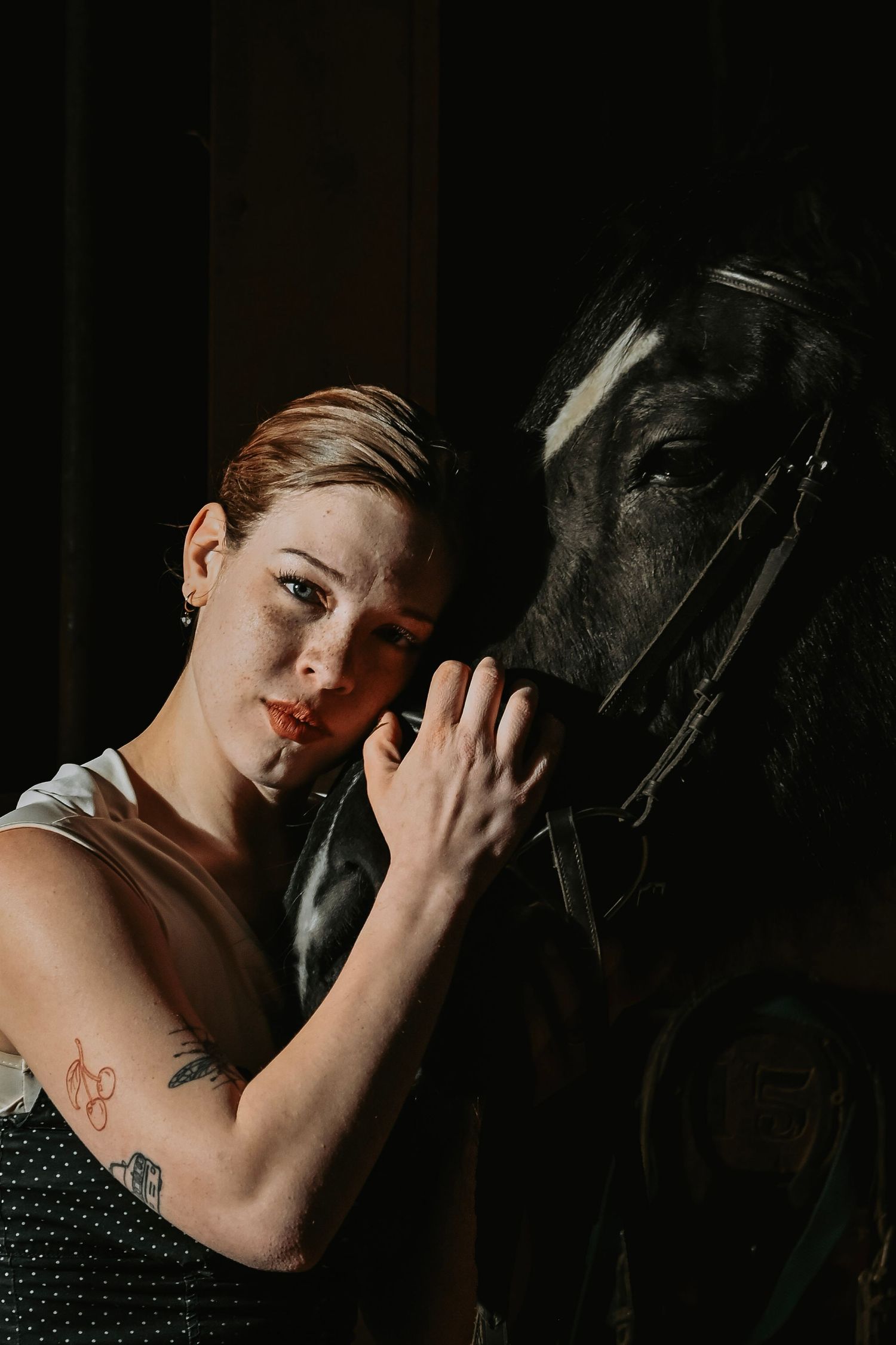 A person in a polka dot dress poses intimately with a dark horse in a dramatic low-light setting.