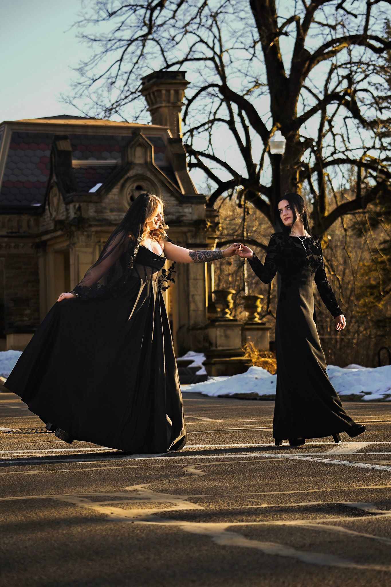 Two figures in elegant black gowns dance gracefully in front of a historic stone building on a winter afternoon.