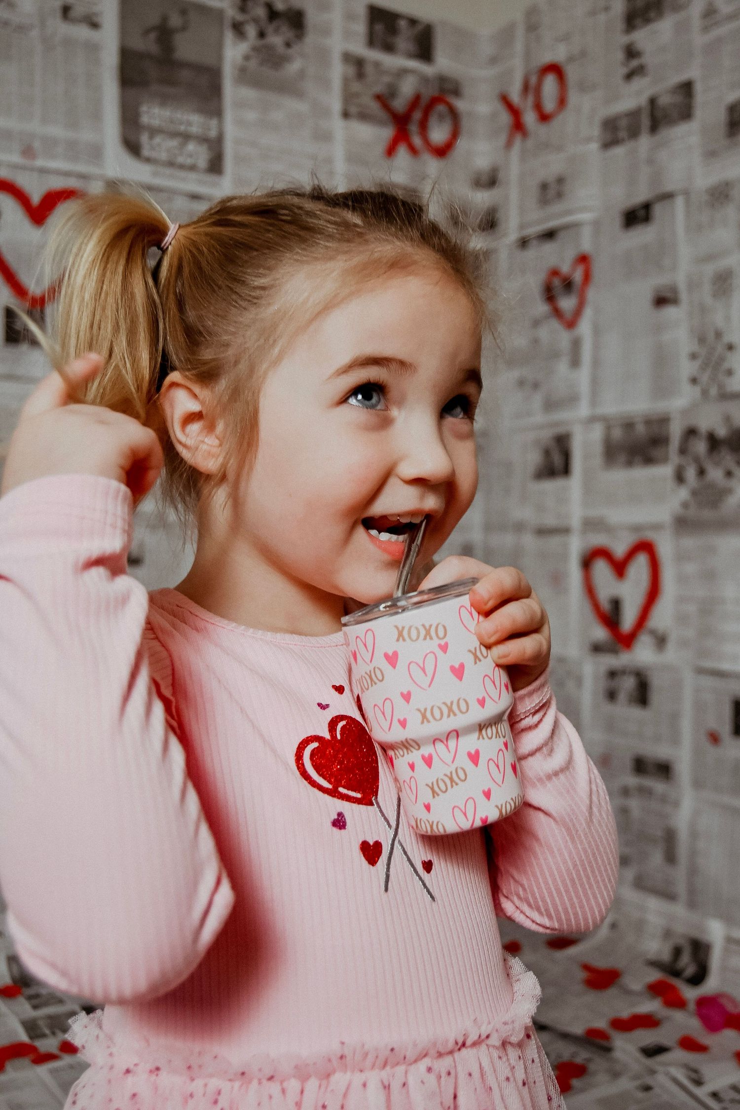 Child in pink sweater holding Valentine cards against newspaper wall with red hearts.