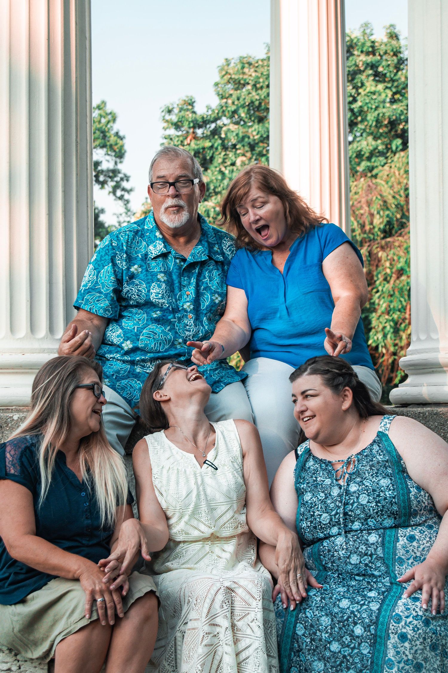 Family group laughing together while sitting on white columns outdoors.