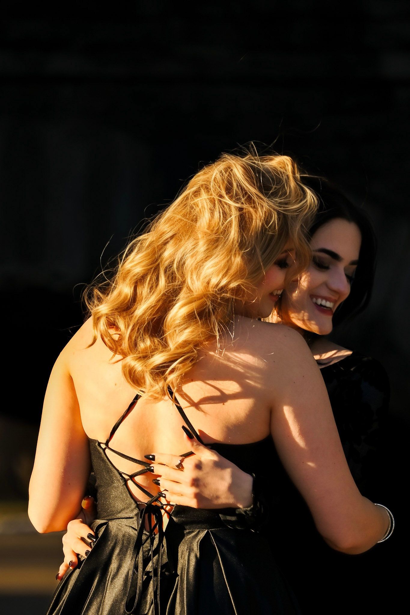 Two women in black dresses embrace and laugh together in warm sunlight casting shadows on their skin.