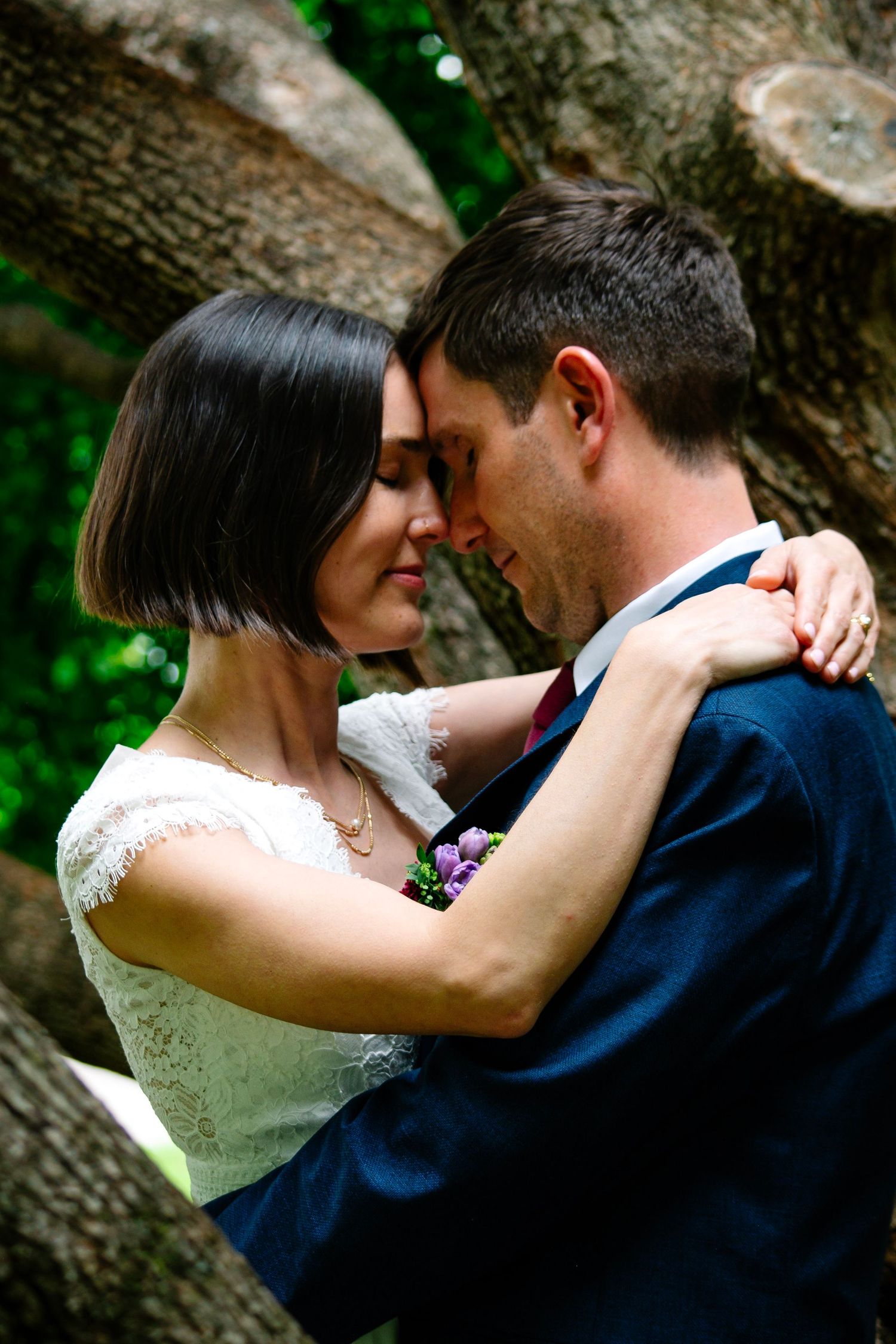 A bride and groom share an intimate forehead touch moment outdoors, surrounded by lush green trees on their wedding day.
