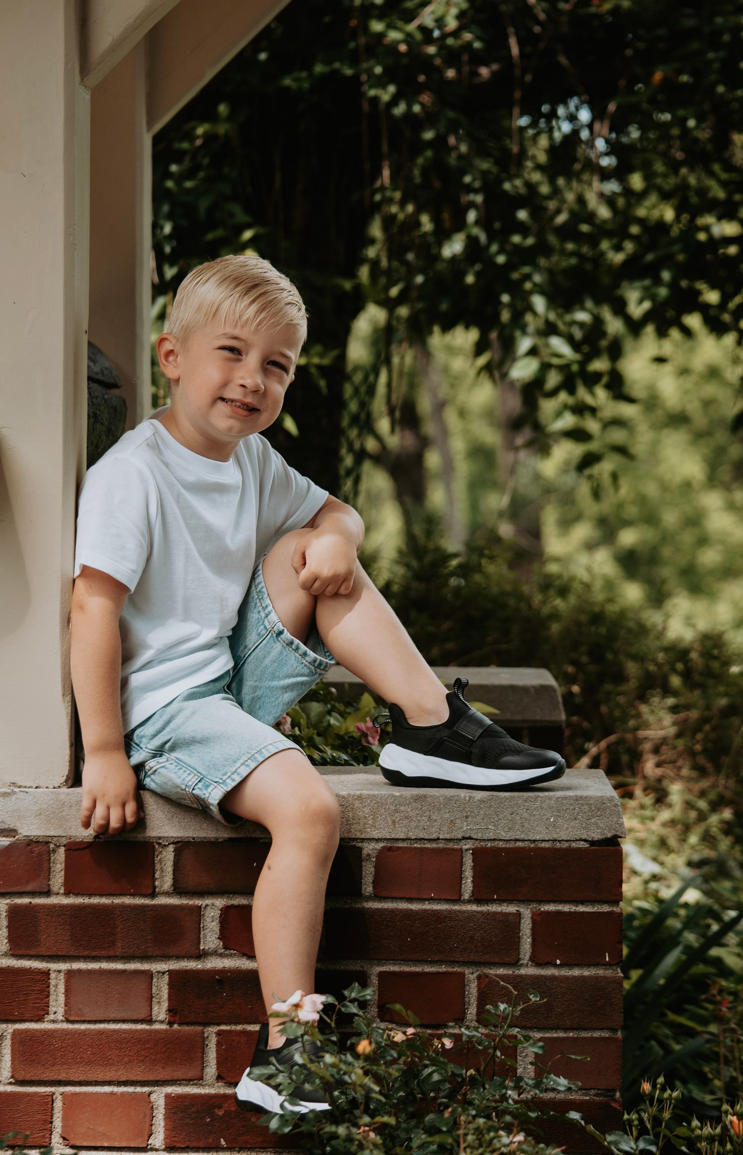 Smiling blonde boy in white tee and denim shorts sitting on a brick wall outdoors wearing black sneakers.