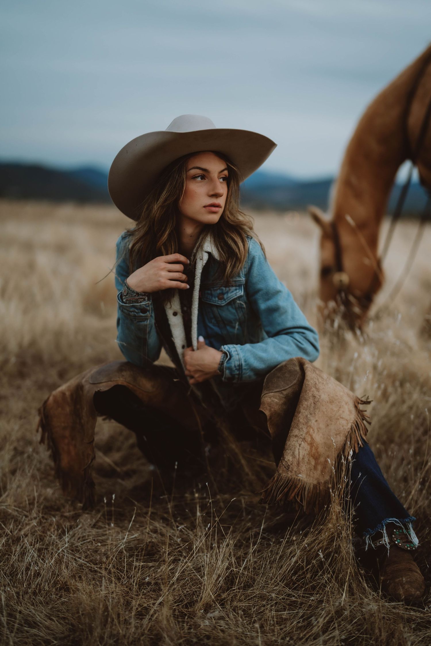 Western Cowgirl in denim jacket and cowboy hat resting in dry grass field with horses.