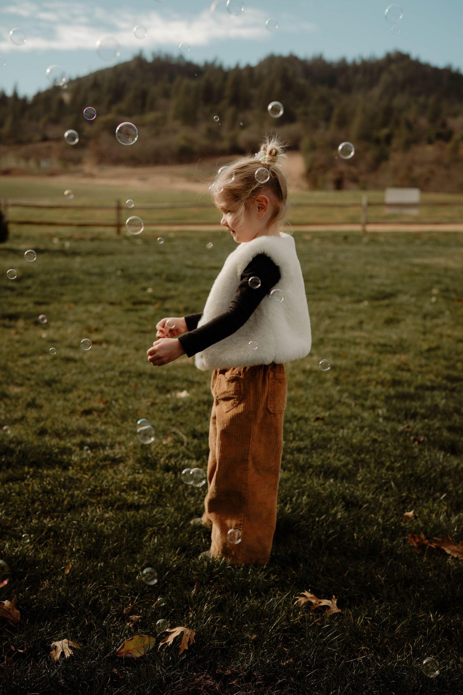 Child in white sweater and brown overalls chasing bubbles in a sunny field.