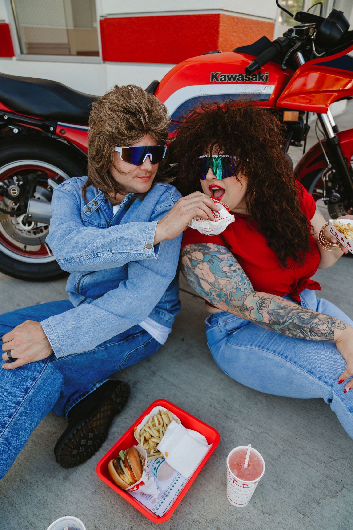Couple relaxing on ground next to red Kawasaki motorcycle with In-N-Out food.