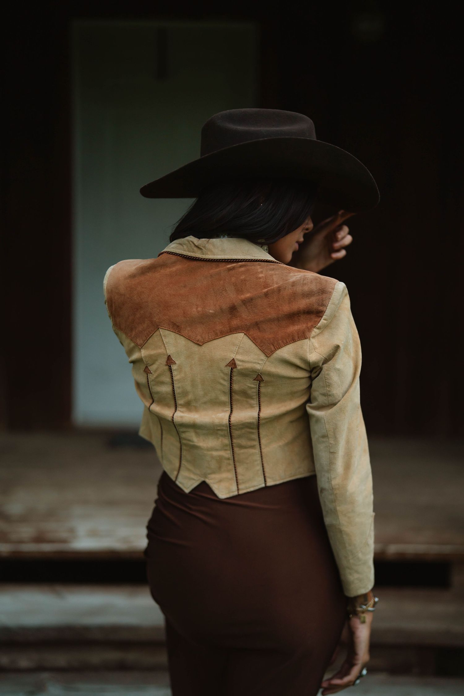 A person wearing a beige and brown western-style outfit shown from behind in dramatic lighting.