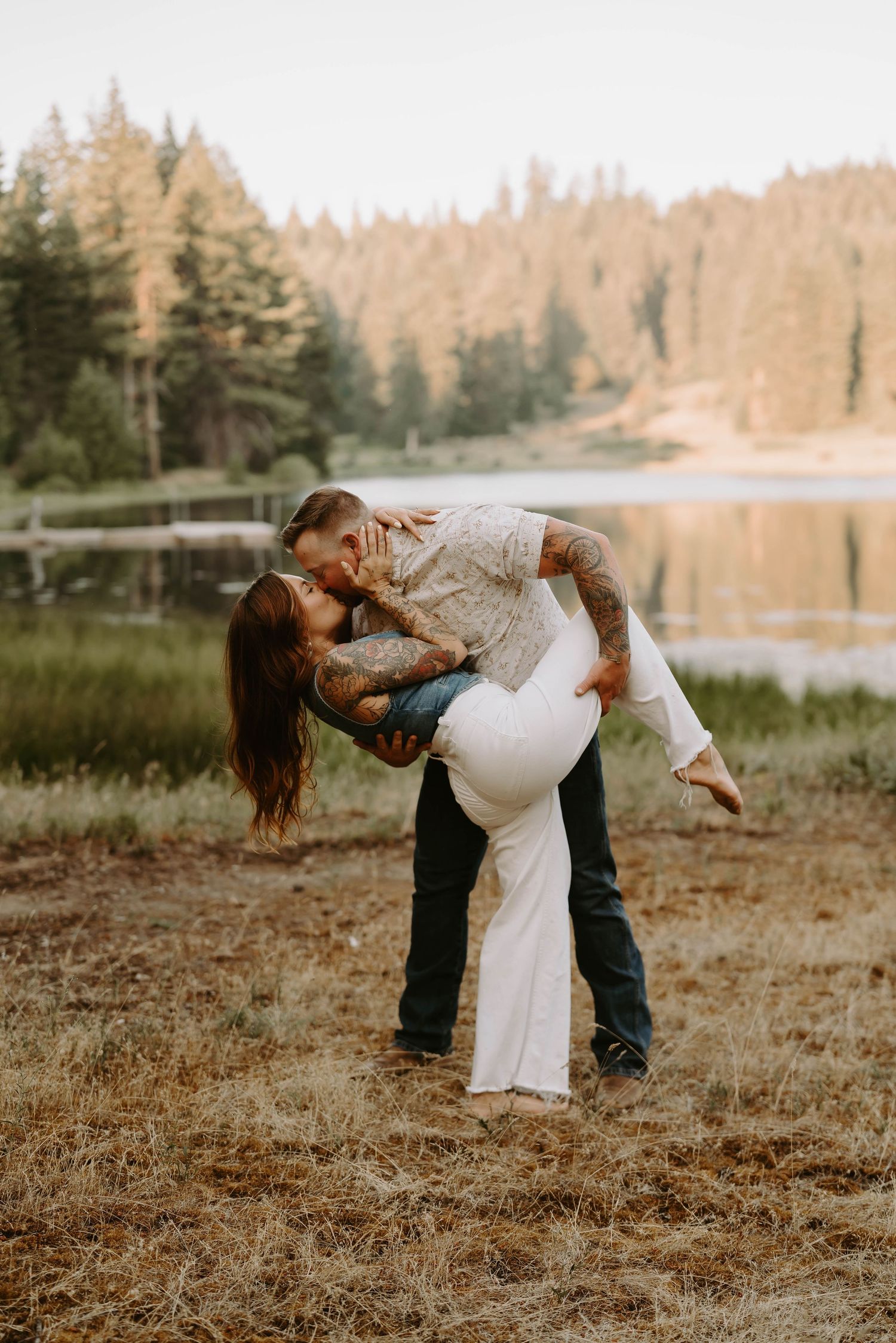 A romantic couple dances playfully in a field near a lake with pine trees at sunset.