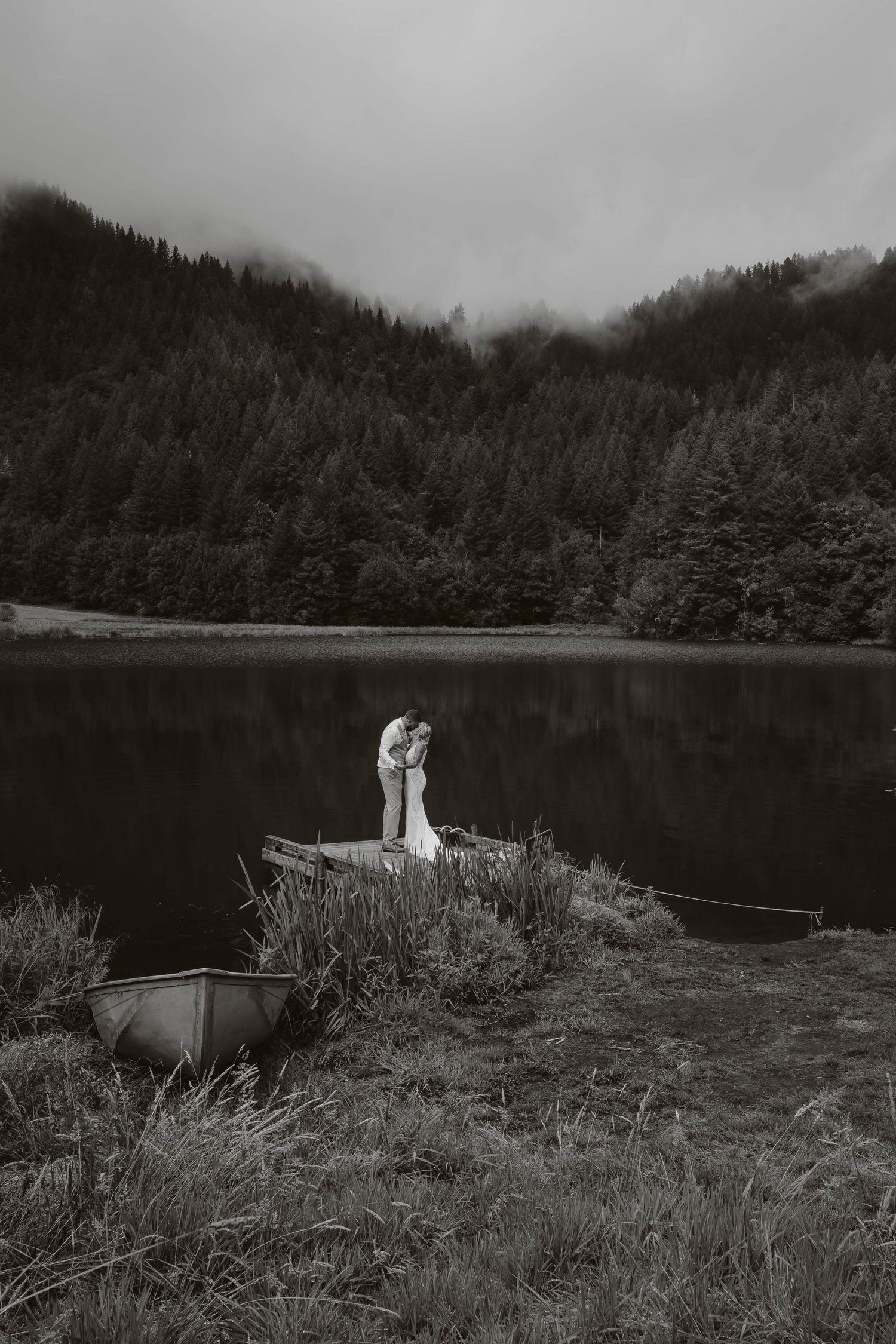 A black and white wedding photo captured by a serene lake surrounded by misty mountains.
