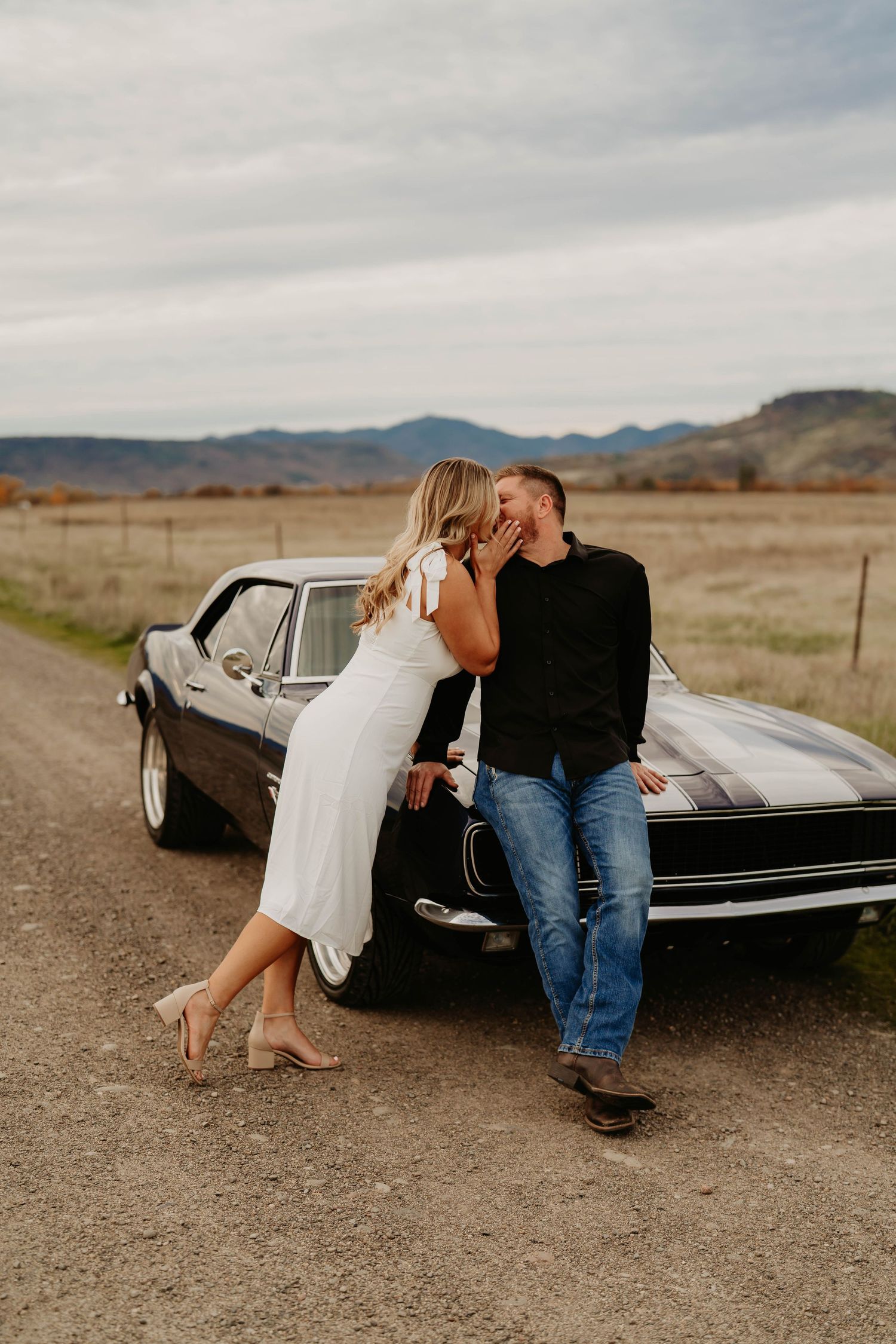 Romantic couple poses with vintage black car in desert setting.