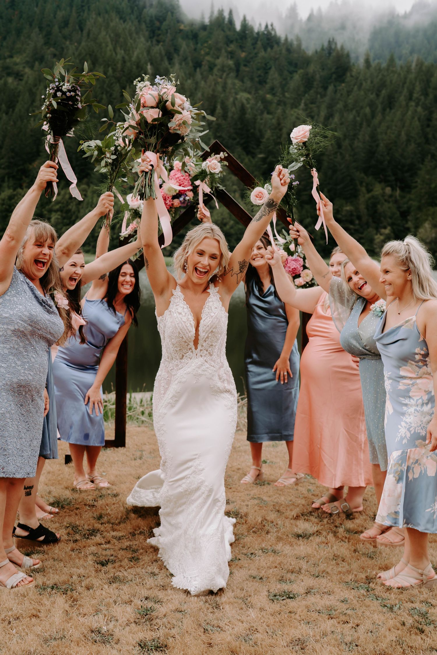 Wedding party celebrating with arms raised on mountainside in pastel dresses.