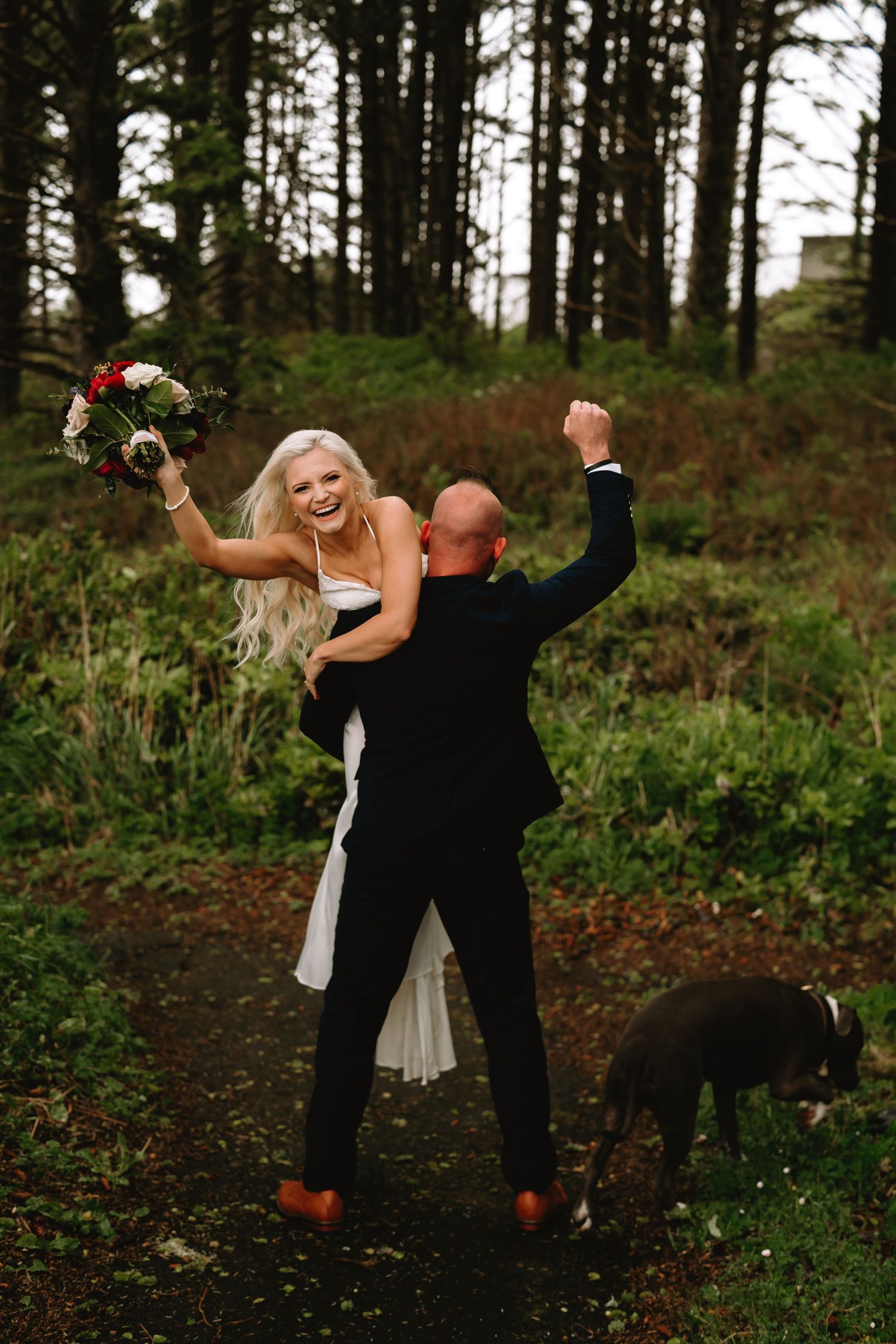 Playful wedding couple dancing in a forest with their dog.