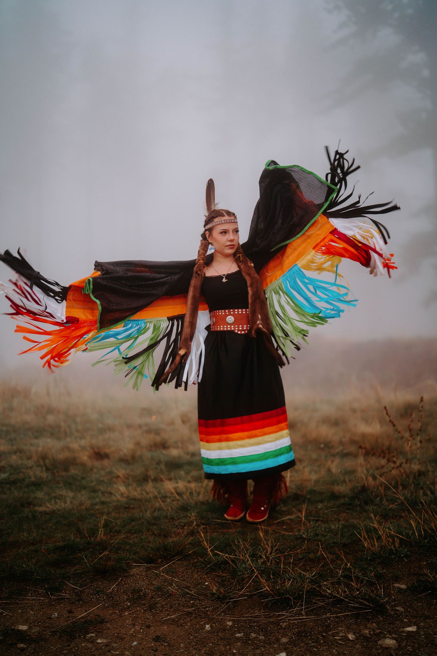Traditional dancer in colorful regalia performs against foggy natural backdrop.