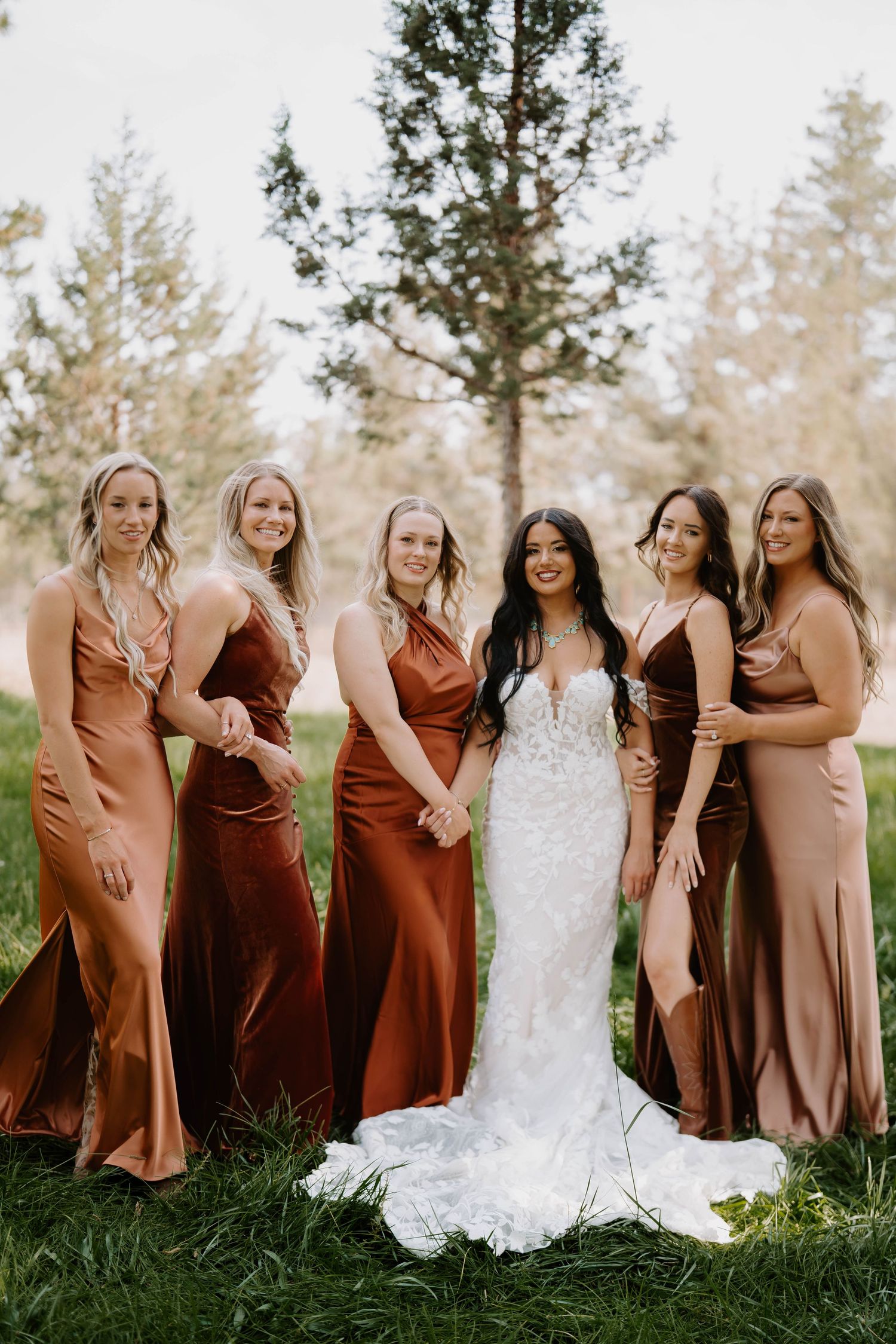 Bridesmaids in rust-colored dresses pose with bride in white lace gown against a natural outdoor backdrop.