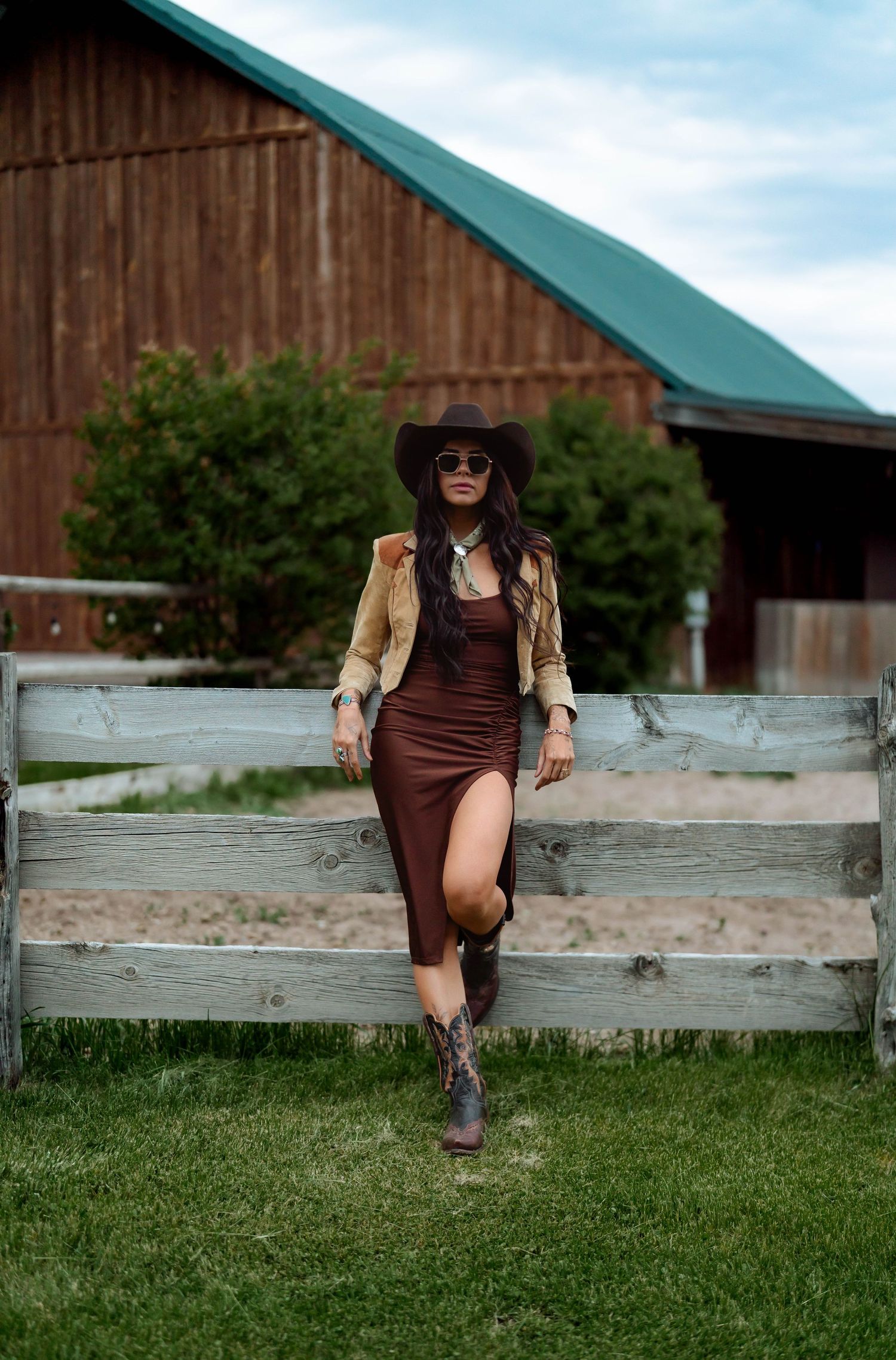 Country fashion scene with brown dress against weathered fence and green-roofed barn.