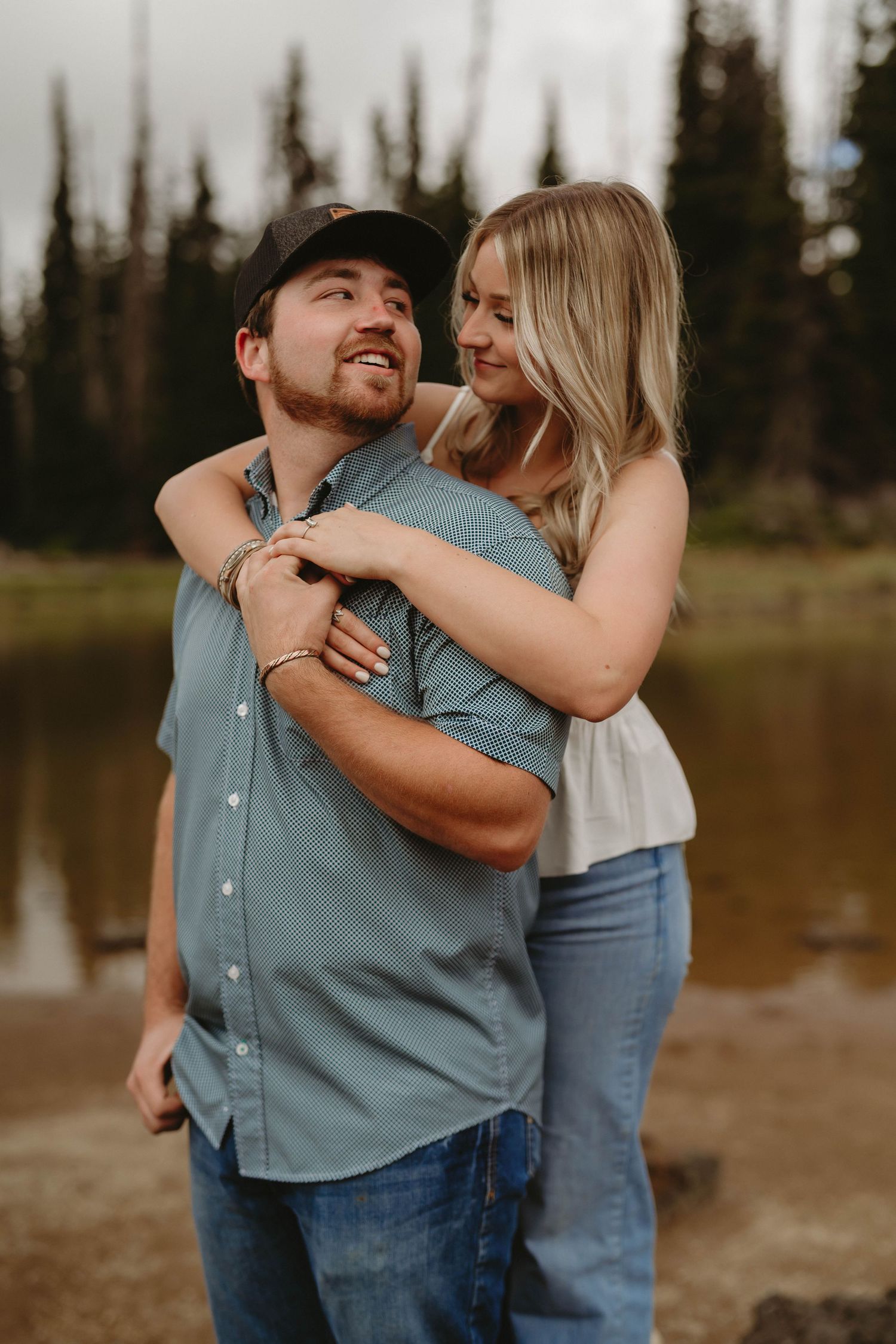 Playful couple embracing outdoors with mountains and forest in the background.