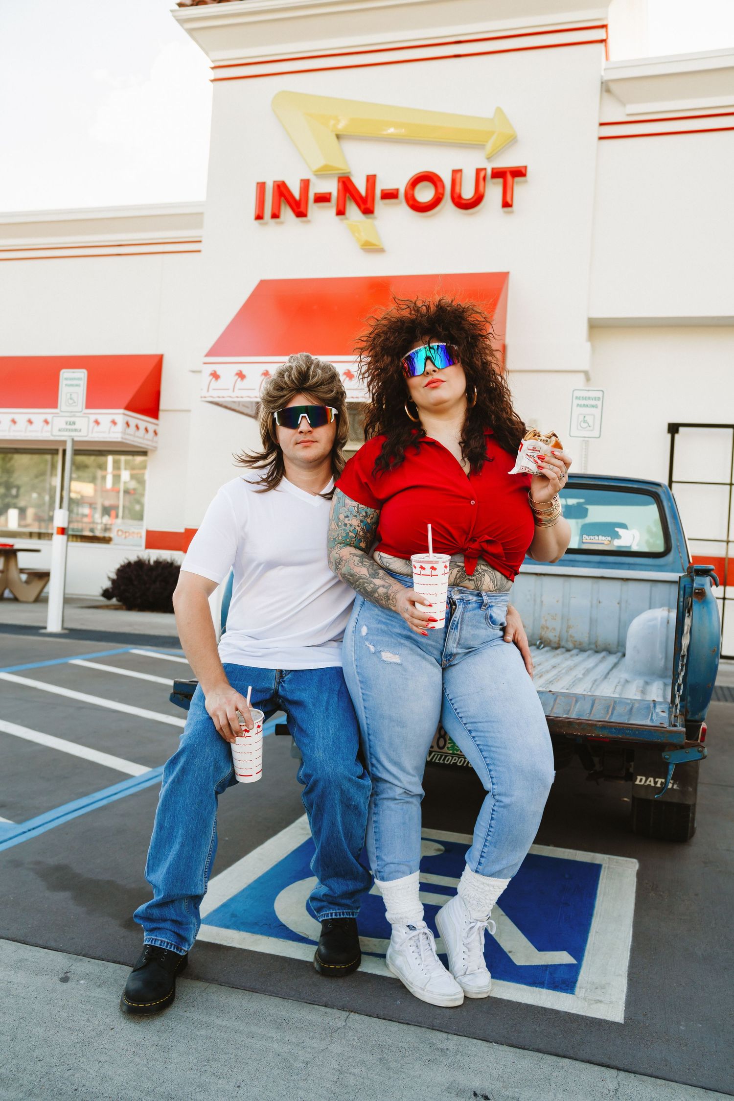 Playful couple in casual wear posing outside In-N-Out restaurant with drinks.
