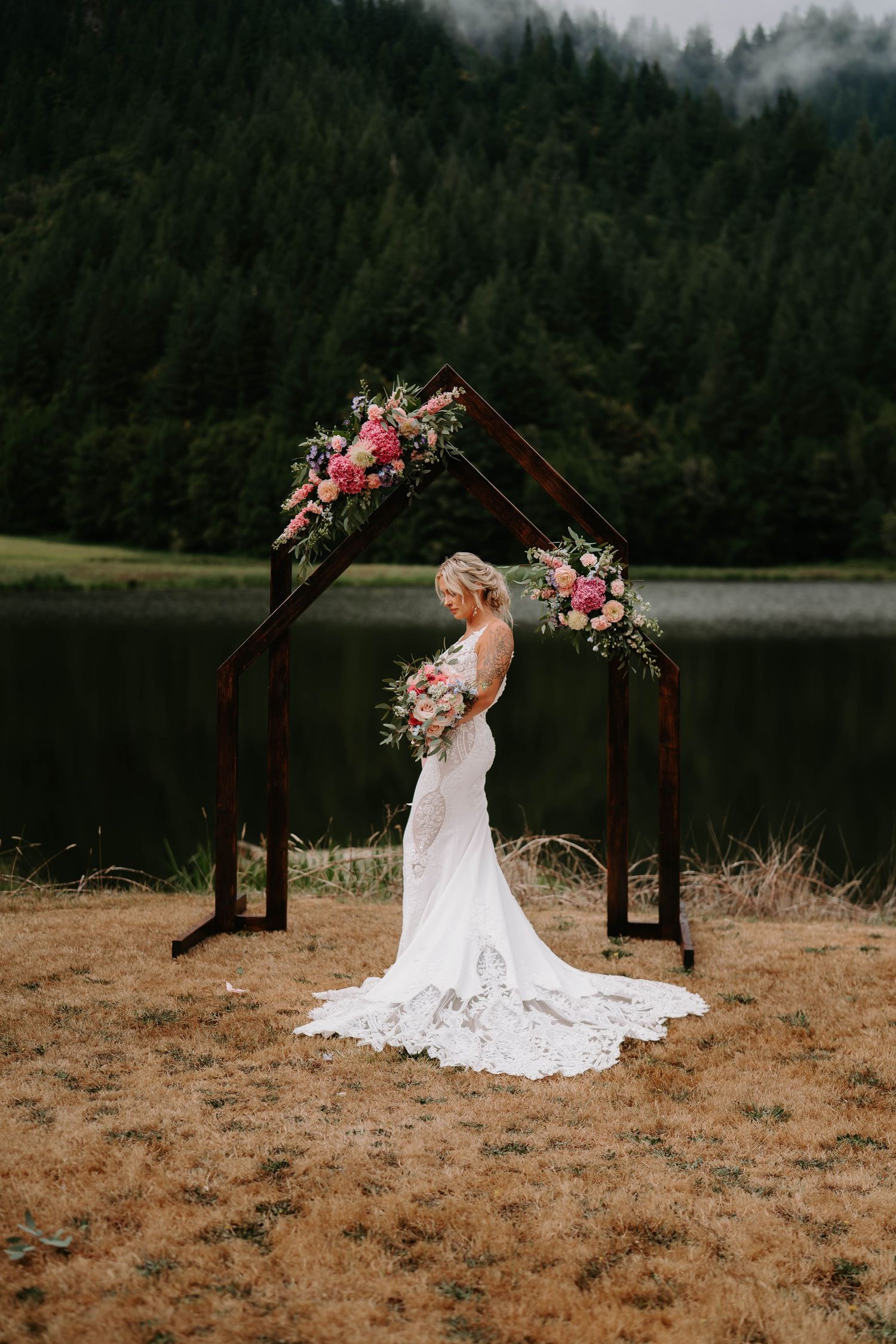 A romantic wedding arch decorated with pink flowers stands against a misty forest lake backdrop.