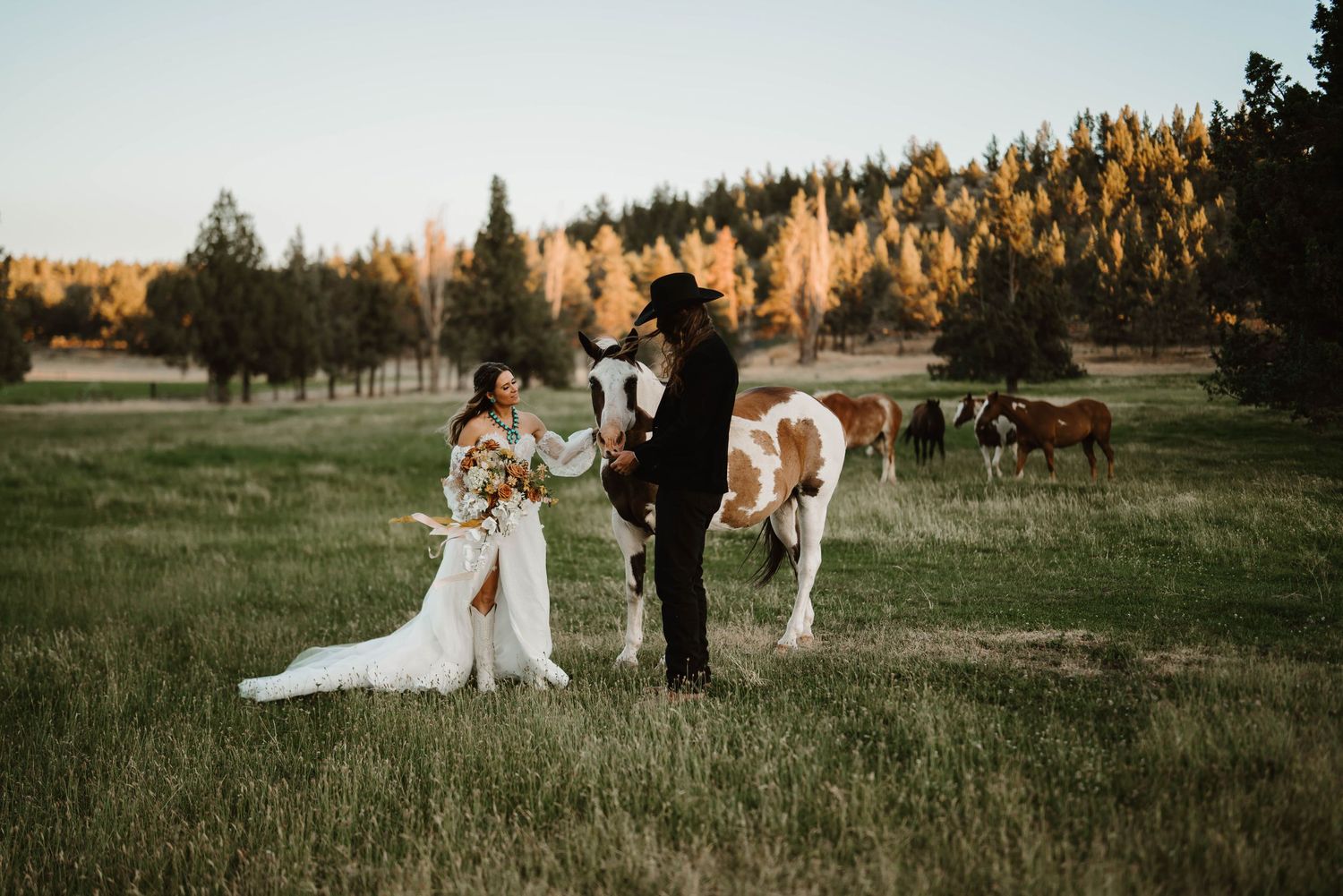 Wedding couple in a pastoral setting with horses grazing in the background at sunset.