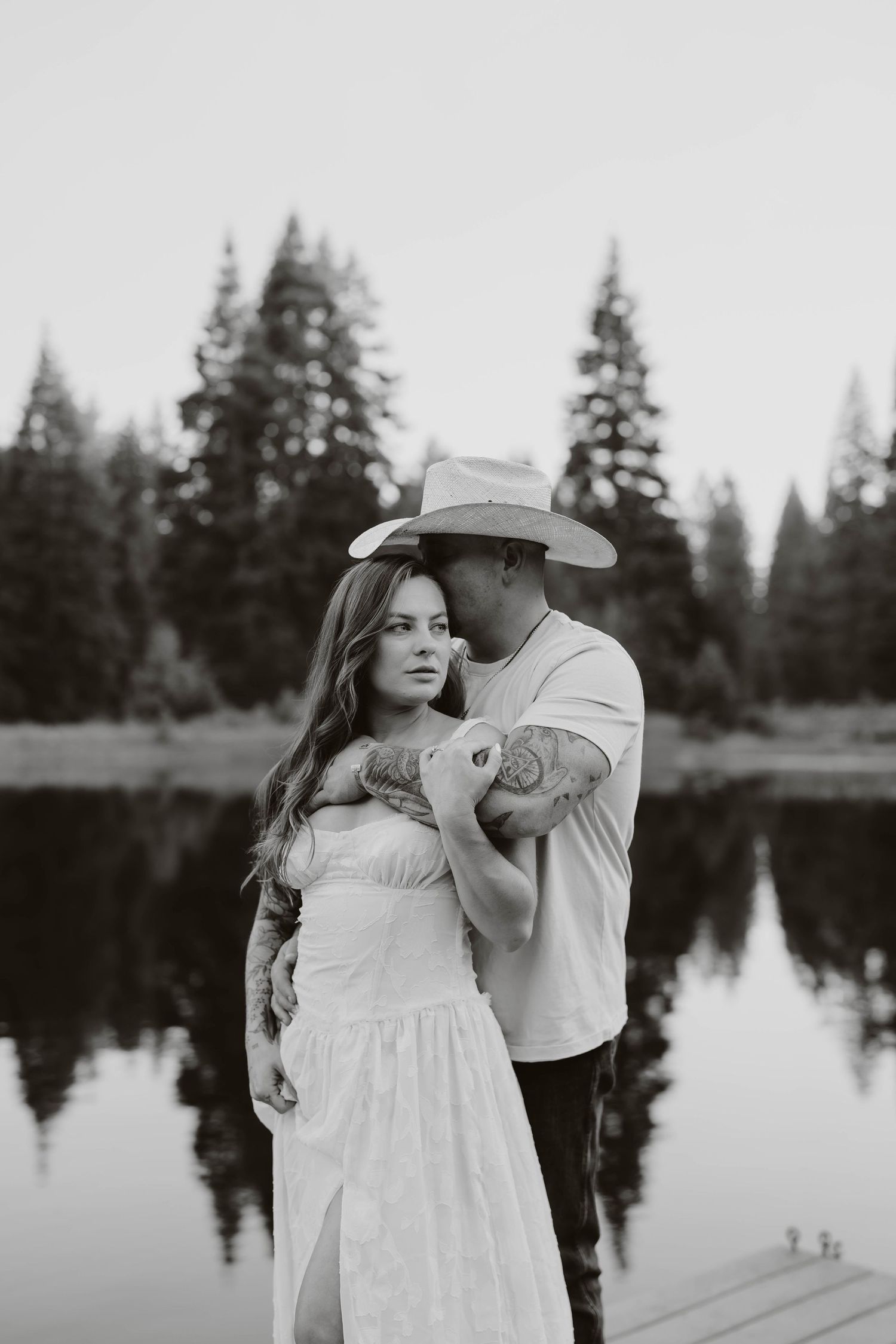 A romantic black and white portrait of a couple by a lake with pine trees in the background.