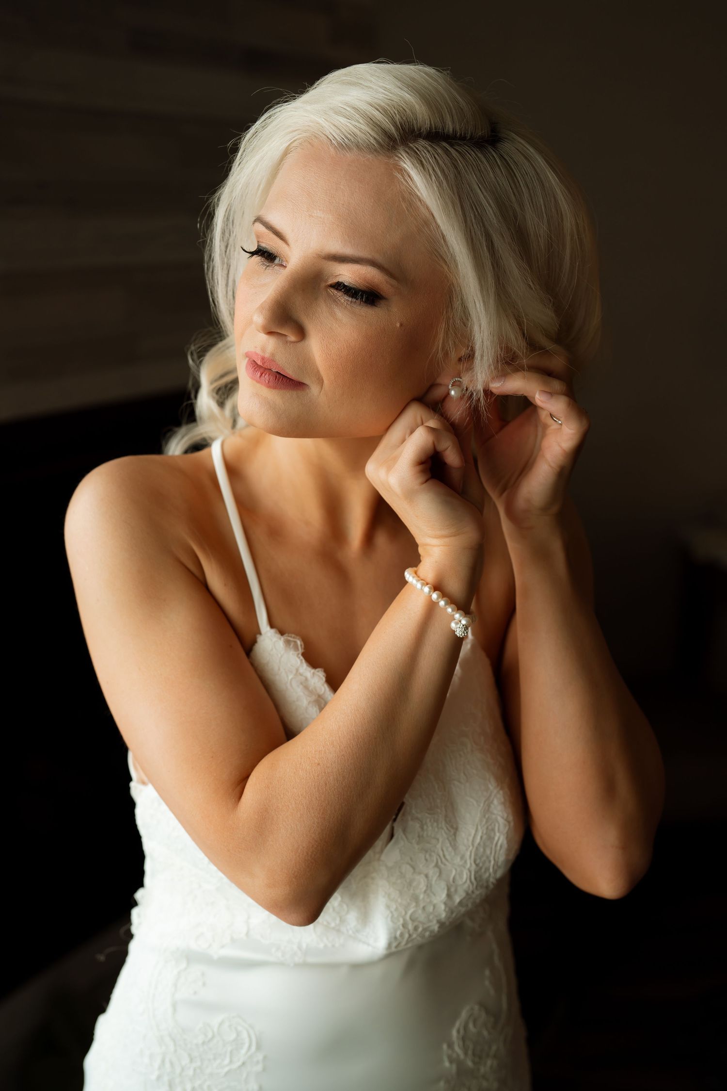 Close-up of a bride in a white lace dress putting on earrings during wedding preparations.