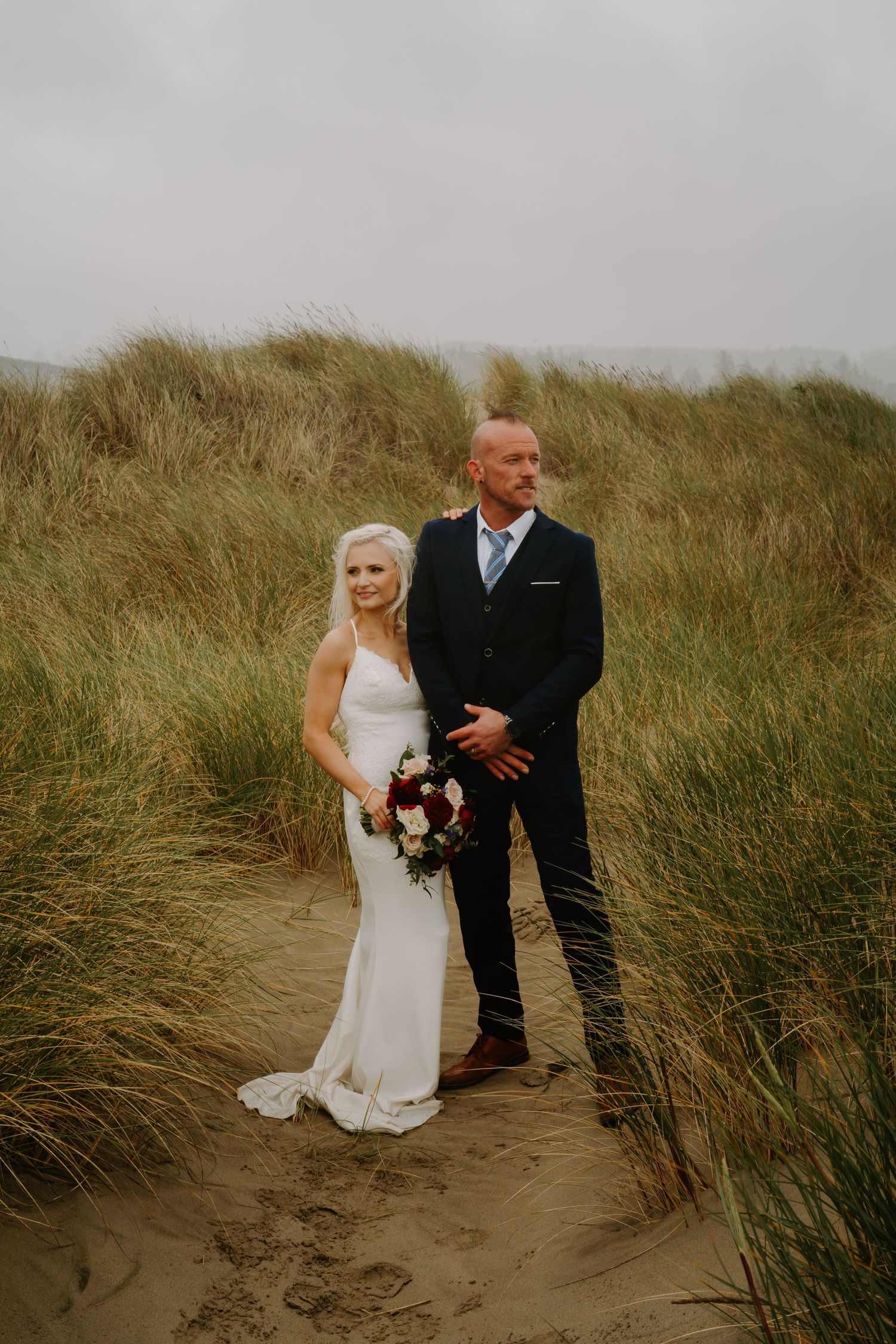 A couple walks through beach grass during their coastal wedding photoshoot.