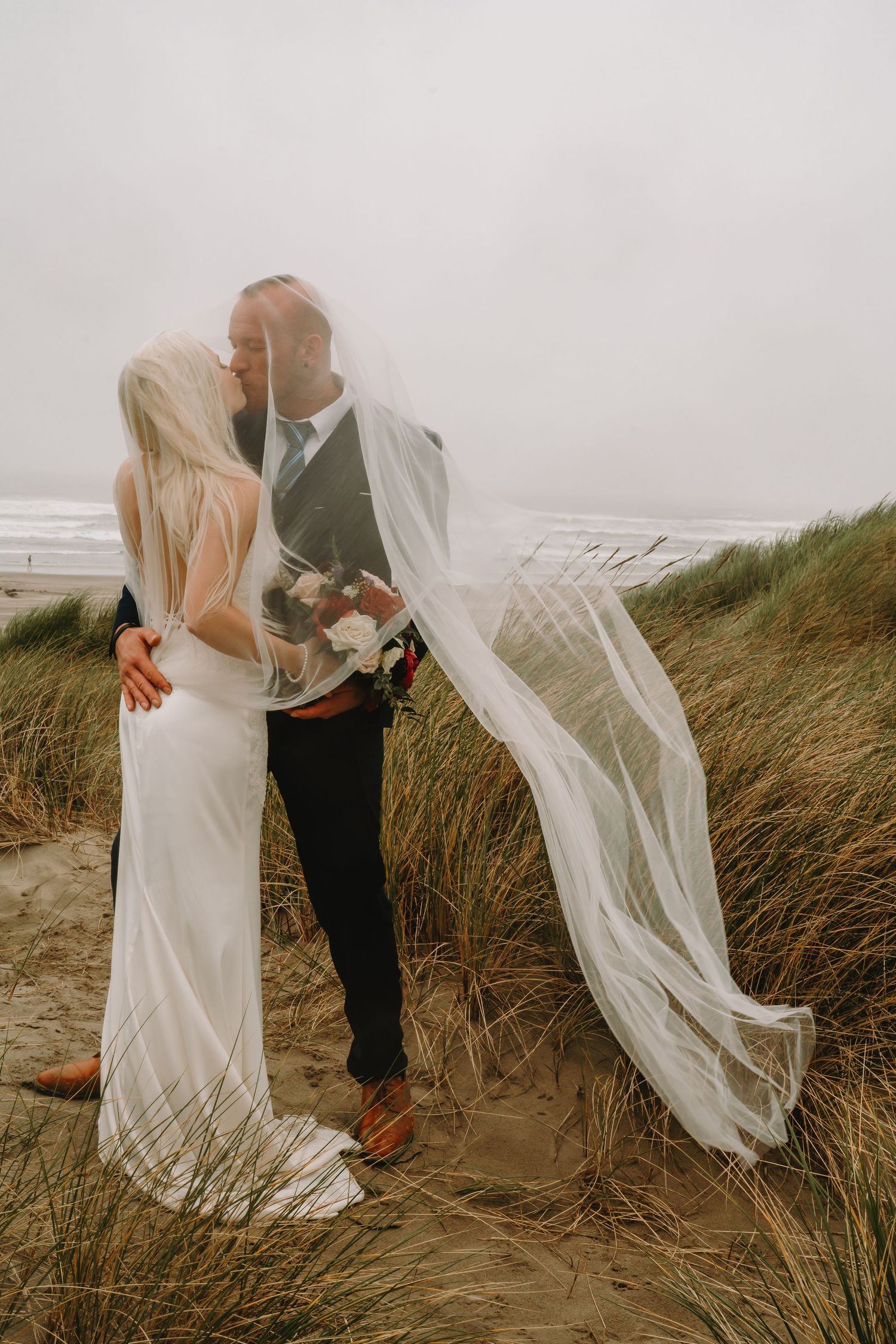 A wedding couple embraces as a veil flows in the wind on a beach with tall grass.