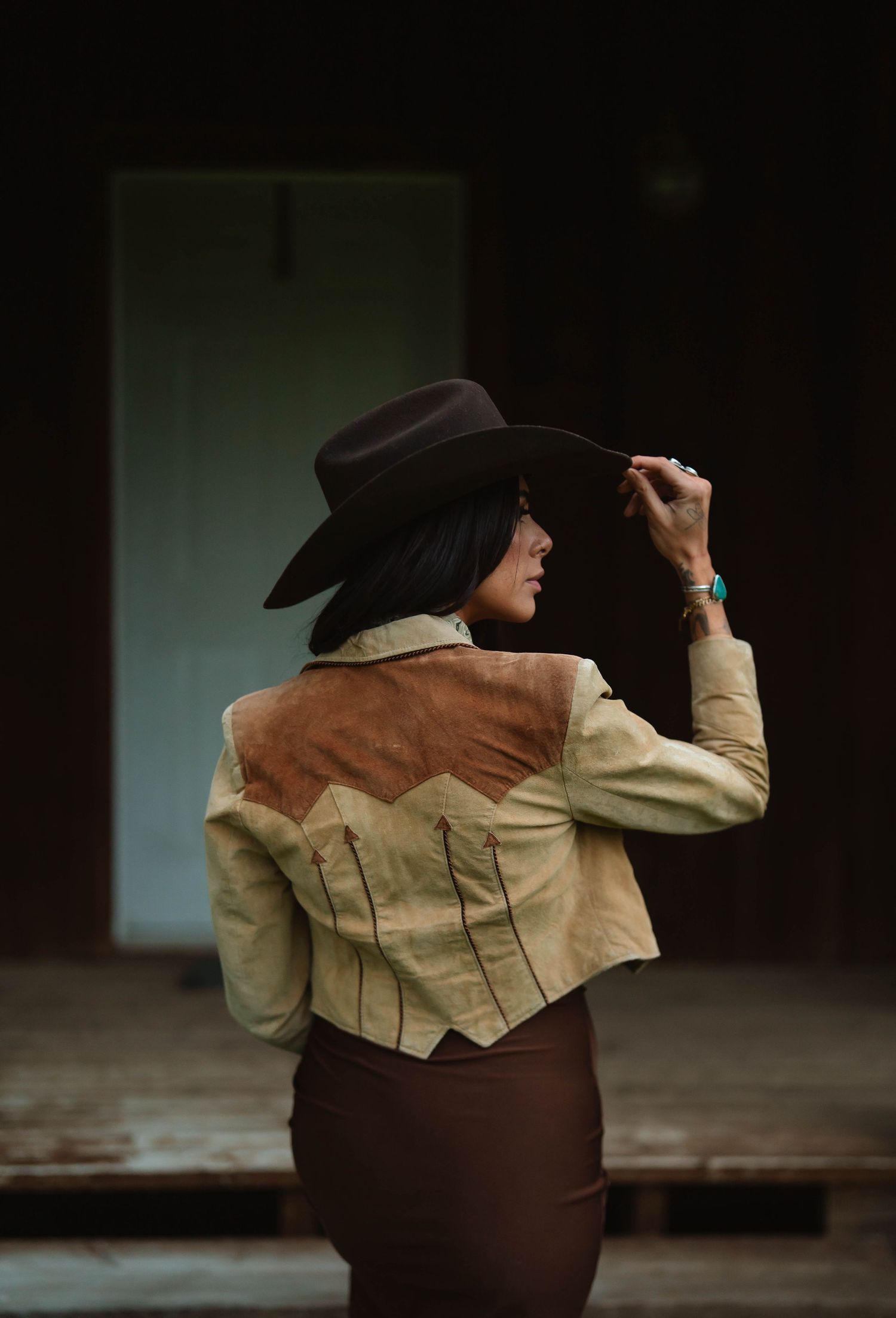 A figure in vintage western attire poses against a dark background in a moody portrait.